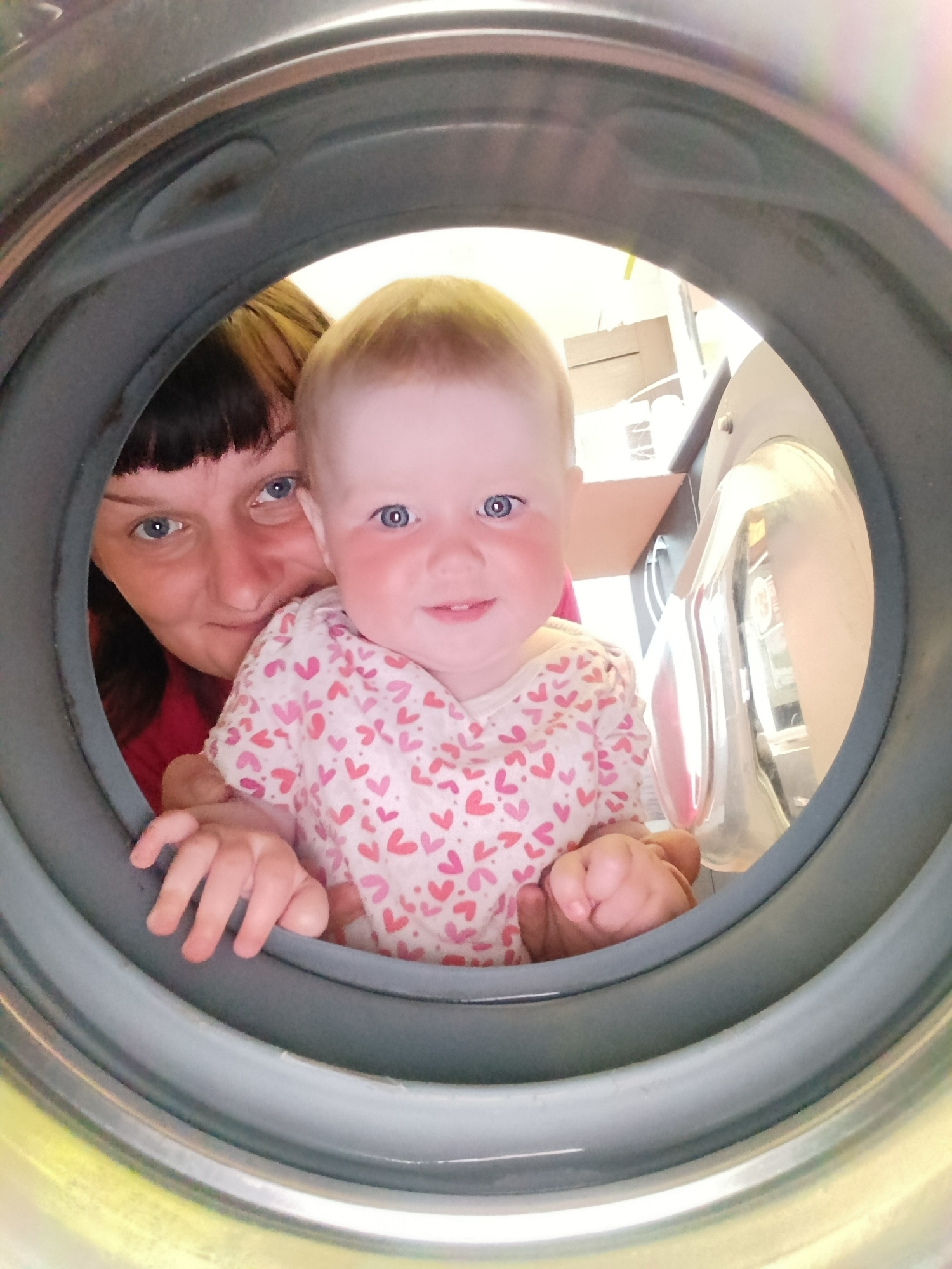 A photo taken through the opening of a washing machine door. The gray rubber seal and edge of the washing machine surround the subjects. Inside the opening are a woman with dark hair and a baby with light hair and blue eyes. The baby is wearing a white onesie with small pink hearts. The woman is looking at the camera with a slight smile. The baby is also looking at the camera, smiling. In the background, partially visible, is a kitchen area with a white cabinet and countertop. The photo gives the impression that the subjects are inside the washing machine, but they are actually looking into it. There is no information to suggest this image was taken in Фоки, Russia.