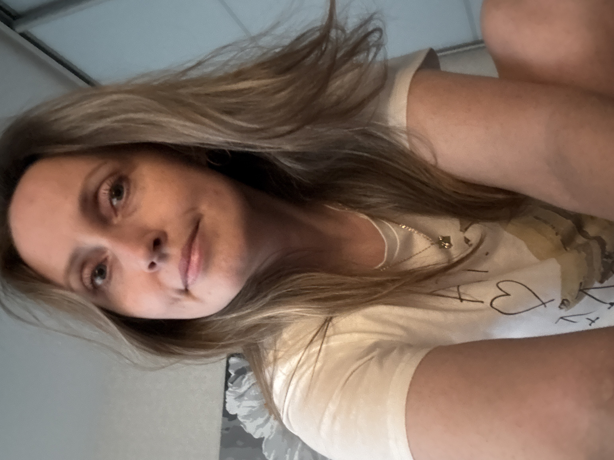 A woman with long, blonde hair is looking at the camera. She is wearing a white t-shirt with a design on it. The background is blurry and indistinct, but appears to show a room. The image is rotated and appears to be taken from a low angle. I am not able to confirm the location.