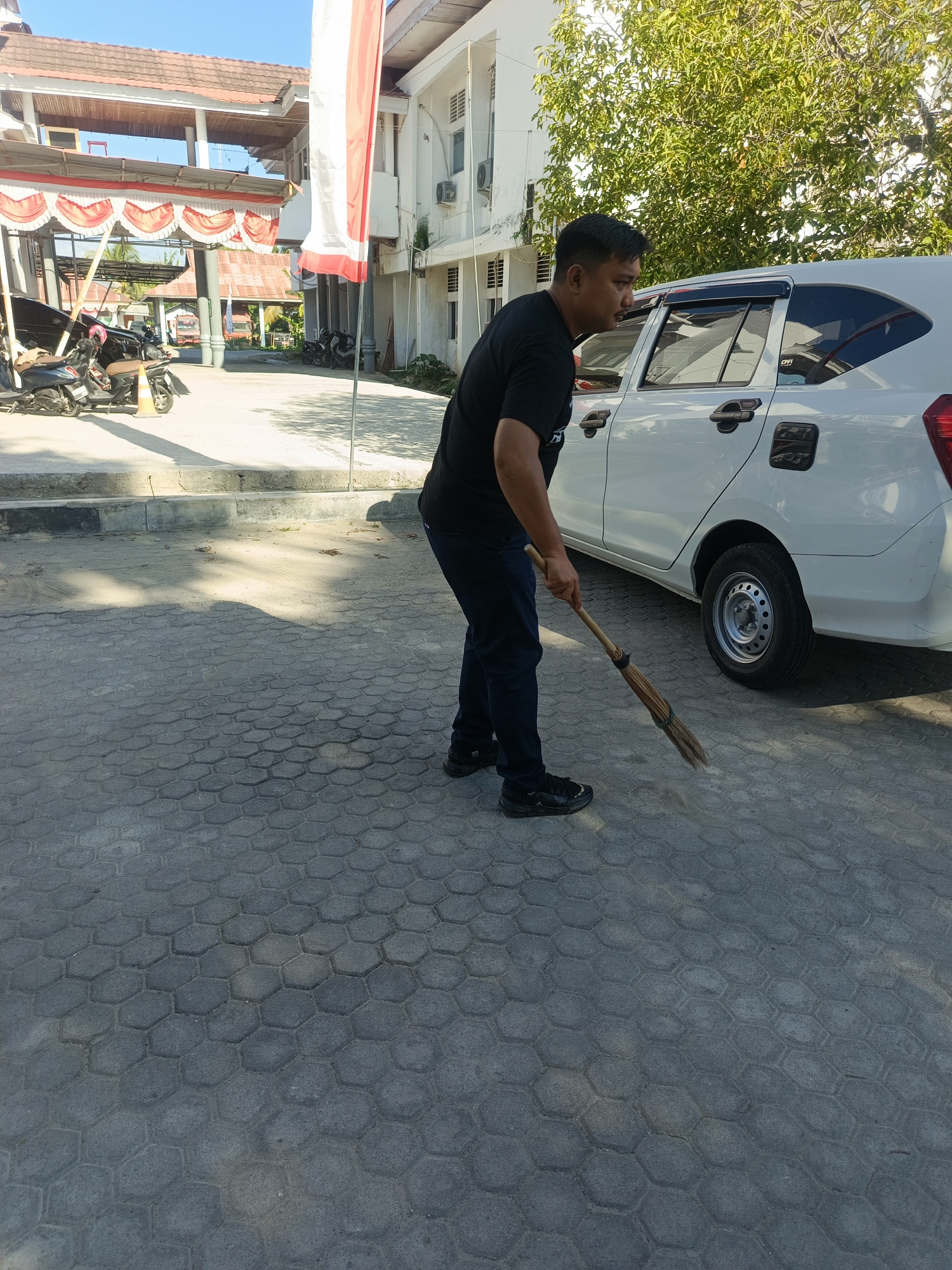 In the city of Gorontalo, Indonesia, a man wearing a black t-shirt and dark pants is sweeping a grey hexagonal paver stone pathway with a broom. He stands to the right of a white car, with the broom in his right hand, actively moving dust. The environment appears to be outdoors, possibly a parking lot or public area, with a building in the background and trees providing dappled shade on the ground. A red and white Indonesian flag is visible on a pole to the left of the building. Several motorcycles are parked on the left side of the image. The sky is clear and blue.