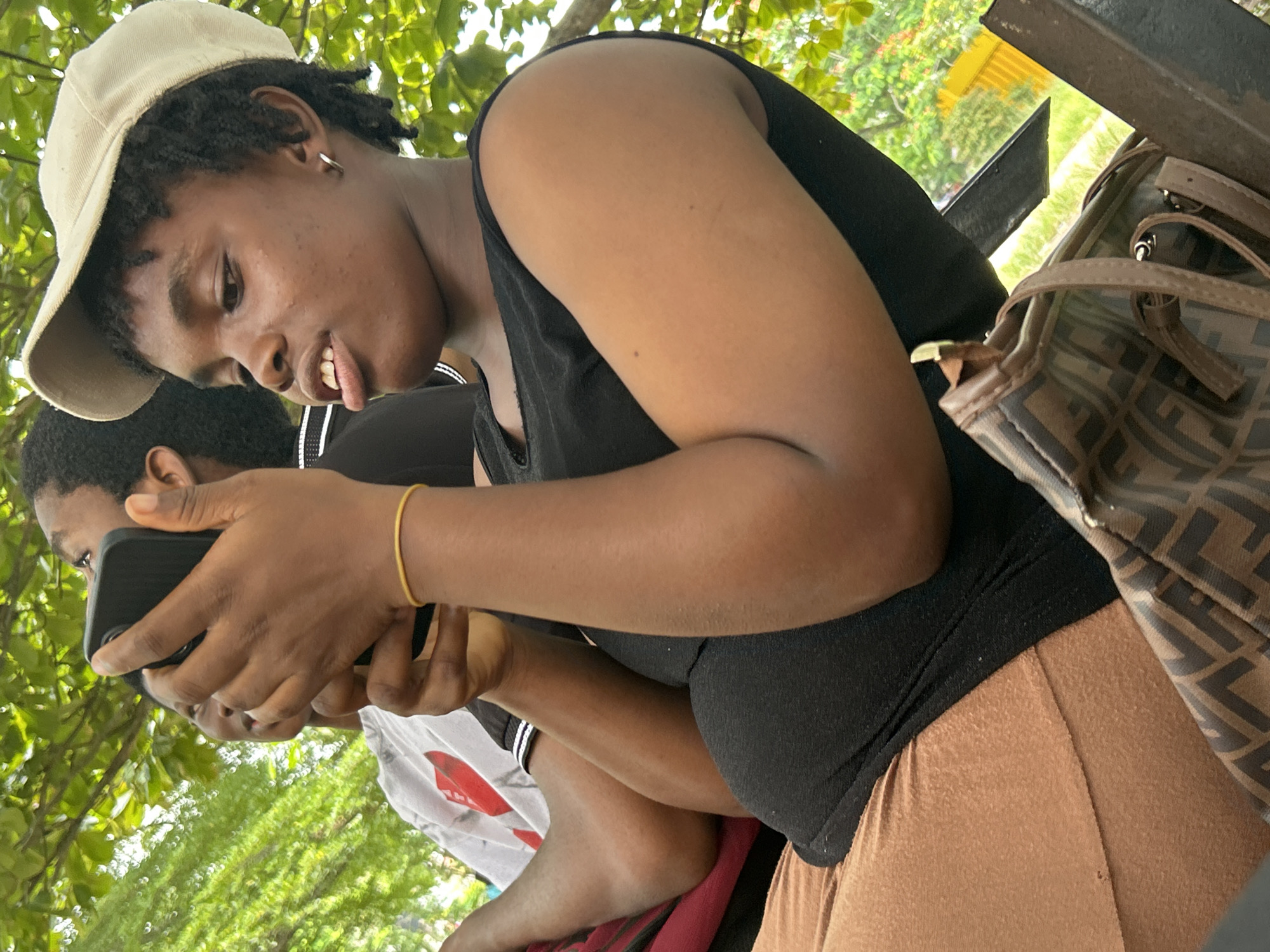 The image shows a close-up of two young people in an outdoor setting, with lush green foliage visible in the background. The person in the foreground, wearing a tan baseball cap, black tank top, and tan pants, is looking down and holding a smartphone. Their tongue is sticking out slightly. A thin gold bracelet adorns their wrist. Partially visible behind them, another person wearing a black shirt with white stripes and a white t-shirt with a red design is also looking down, presumably at the same smartphone. A patterned handbag with brown straps rests on a dark metal surface to the right, partially obscuring the foreground subject's arm. The lighting suggests it is daytime, possibly with overcast or dappled sunlight filtering through the leaves. The overall mood appears casual and relaxed. The location is Ihiagwa, Nigeria. No visible text is present.