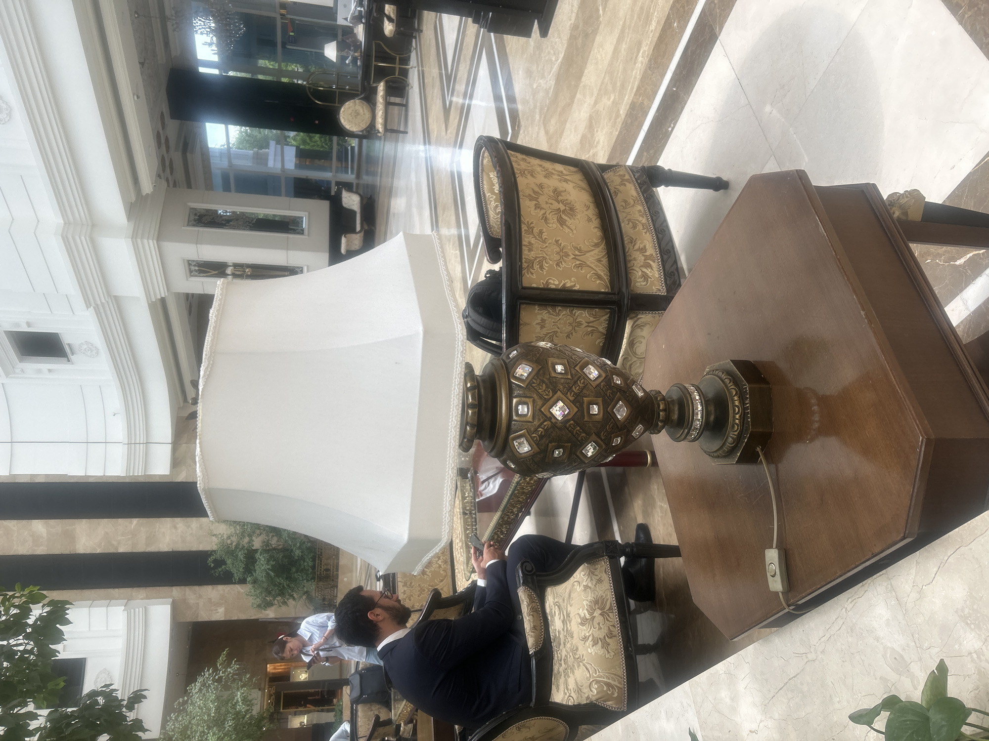 In Almaty, Kazakhstan, a man in a dark suit sits in an ornate armchair, looking at his phone. He is in a brightly lit, spacious lobby, likely a hotel or convention center, with marble floors and high ceilings. A large, white lampshade dominates the center of the frame, with a decorative, crystal-studded lamp on a wooden table in the foreground. Other chairs and furniture are visible in the background, along with a woman in a light blue shirt looking at her phone. The overall impression is one of opulence and modern business or leisure.