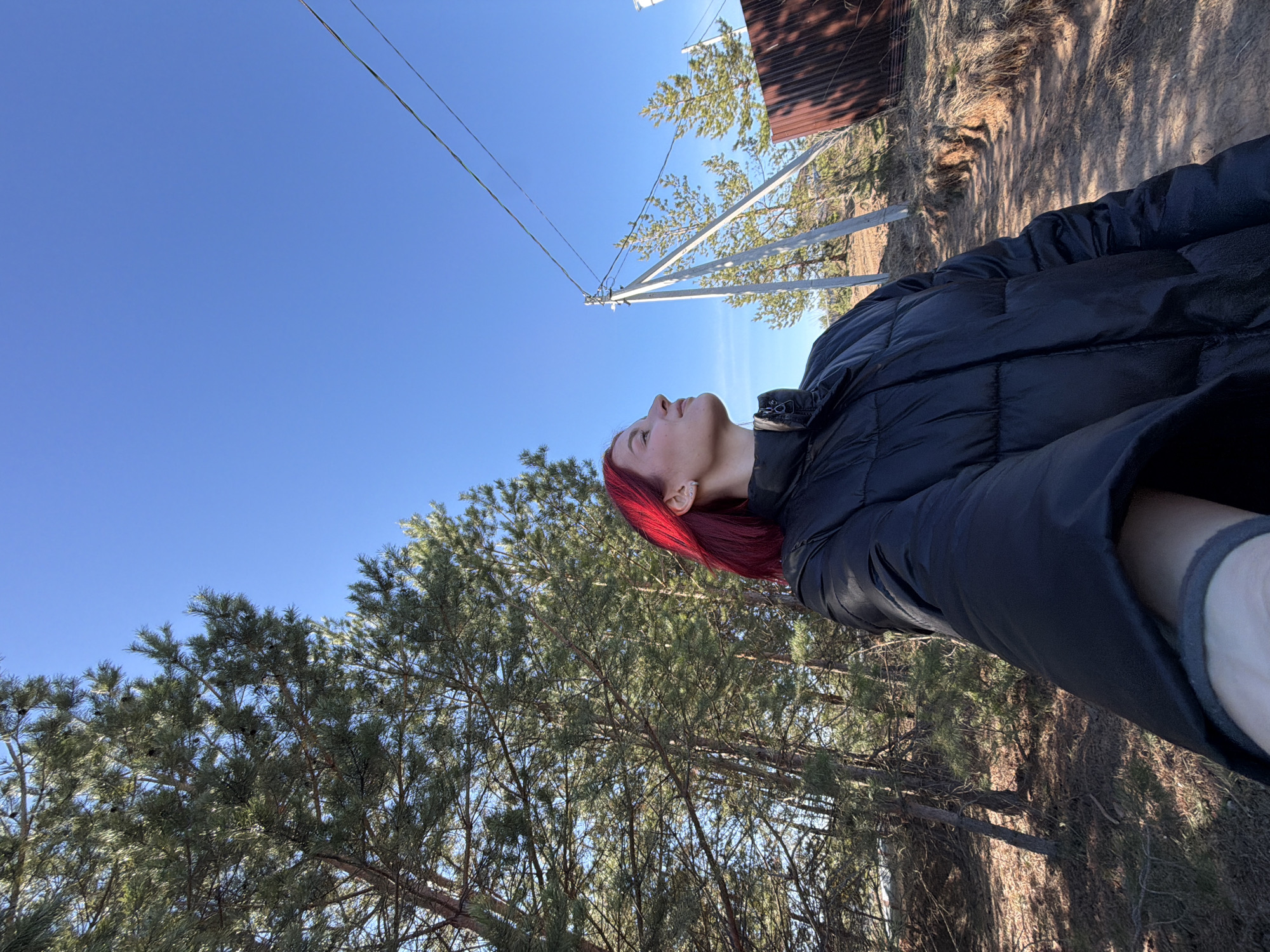A woman with red hair, wearing a black puffer jacket, is looking up at the sky. Above her is a clear blue sky with utility lines. Behind her are trees, a wooden structure, and a fence. The photograph appears to be taken outside in a natural setting. It is likely located in Chaikovsky, Russia, but there is nothing in the image to concretely support or deny this.