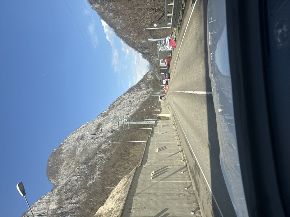 The image captures a mountain road in Monastyr', Russia, on a clear, sunny day, viewed from inside a vehicle looking forward. The road is asphalt, two-laned, with white solid and broken lane markings, and appears to be ascending or winding through a valley. Steep, rocky mountainsides, sparsely vegetated with leafless trees, flank both sides of the road under a bright blue sky, which features a faint aircraft contrail.
On the left side of the road, a long, segmented concrete retaining wall parallels the road. Multiple streetlights on tall poles are positioned along this wall. Further down the road, a dark tunnel entrance is visible on the left. Shadows are cast by the wall and roadside objects, indicating direct sunlight from the right.
Ahead, near the tunnel entrance, road construction or maintenance activity is evident. A white truck, possibly equipped with a crane, is positioned in the right lane, followed by a dark sedan. On the right shoulder, a temporary site features various informal structures, possibly sheds or containers. A red vehicle is parked near these structures, and a significantly damaged white vehicle, possibly a car with "AMG" visible on its side, rests precariously against some of the temporary structures and red barricades. A metal guardrail runs along the right edge of the road, and a white triangular "give way" sign with a red border is visible. A distant, snow-capped mountain peak can be seen through the valley opening. No individuals are distinctly visible.