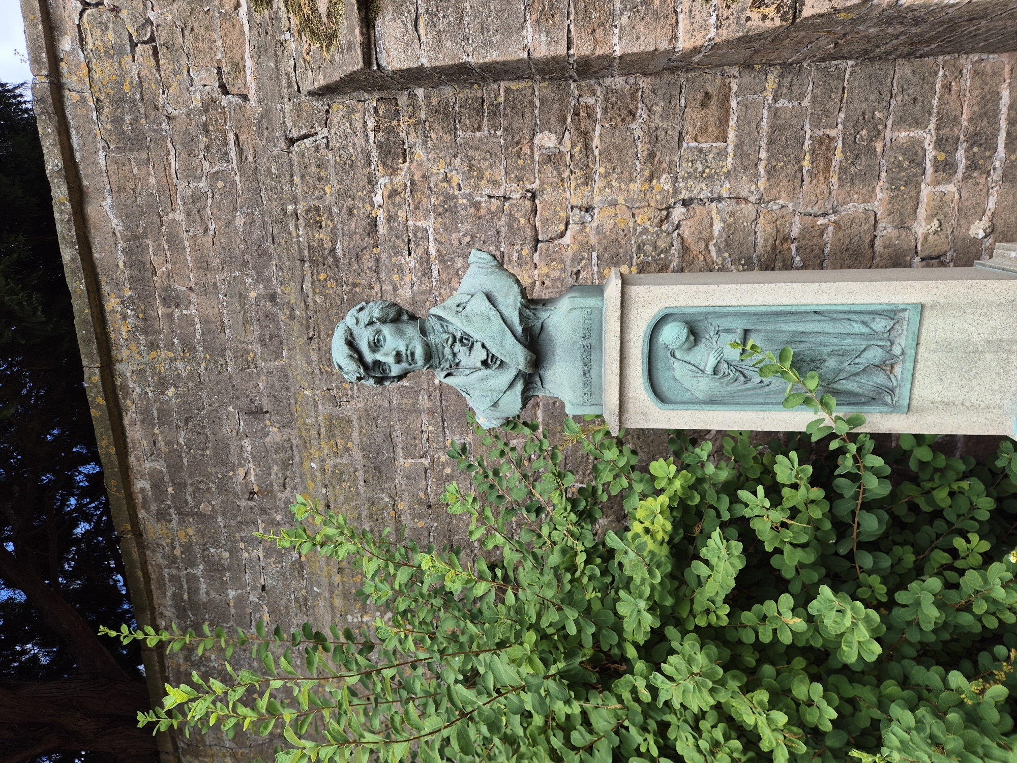 An outdoor, eye-level shot shows a bronze bust of a man, identified by an inscription as "GILBERT WHITE", atop a weathered stone pedestal. The bust is positioned to the right of a lush green shrub, with its branches reaching towards the pedestal. Behind the bust and shrub is a rough, textured stone wall, part of a building or structure. The wall has irregular stones and visible mortar, with some green moss and yellow lichen growth. A dark tree is visible in the upper left corner, with dark branches and green foliage against a blue sky. The pedestal itself has a relief carving of a robed figure holding a laurel branch on its front face. The image appears to be taken in Gedling, United Kingdom, likely at a historical site or memorial.