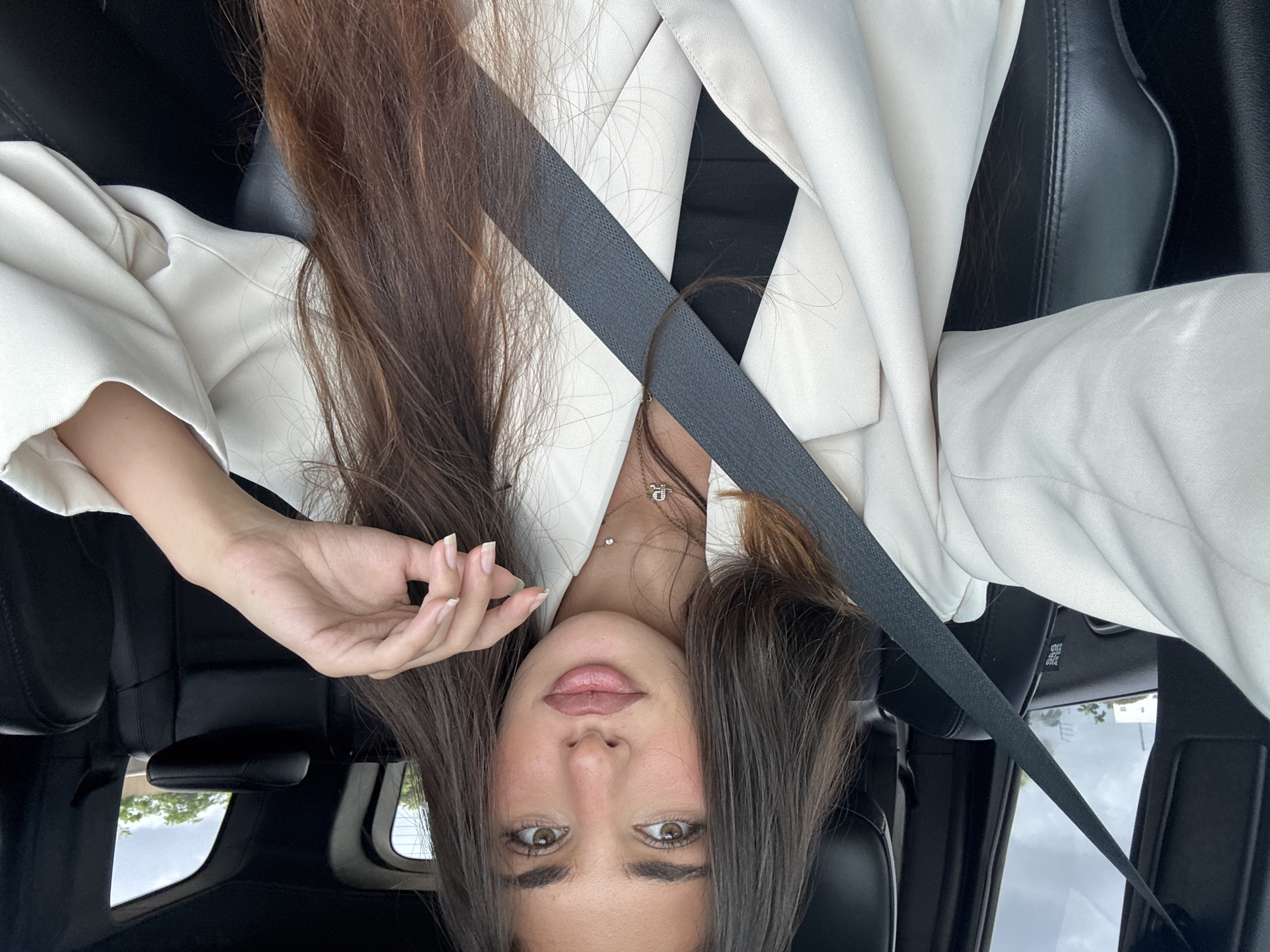 A woman is shown in an upside-down orientation inside a car with black leather seats. She wears a white blazer and a black top. She has brown hair and is wearing a seatbelt. A view through the car window shows trees and buildings, indicating an urban or suburban environment. The image was taken in North Miami, United States of America.