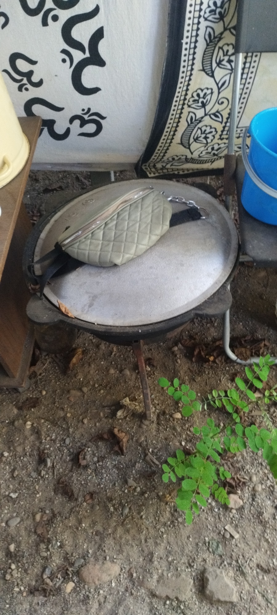 A light-colored, quilted fanny pack rests on top of a large, dark, circular cooking pot that is supported by three legs. The pot is situated outdoors on a dirt and gravel ground. To the left of the pot, a portion of a wooden shelf with a beige plastic bucket on it is visible. Behind the pot, a wall is adorned with black Om symbols and a patterned fabric hangs down the right side. A bright blue bucket stands to the right of the pot. In the foreground, green plants are growing out of the ground. The scene appears to be in an outdoor setting, possibly a backyard or patio area.