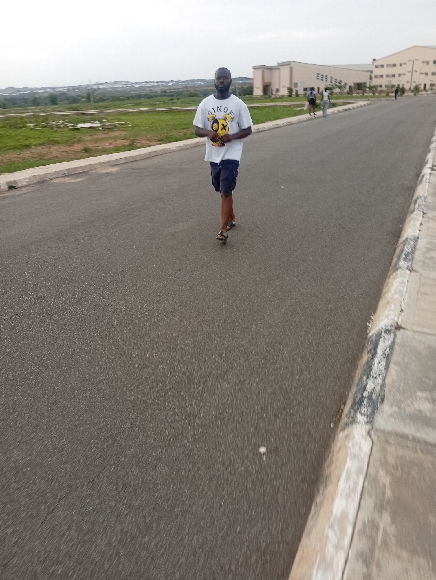 A man walks along an asphalt road in Yelwa, Nigeria. He is wearing a white t-shirt with a yellow and black smiley face graphic and dark shorts. The road is bordered by a concrete curb and a grassy area. In the background, there are buildings, some with windows and architectural details, and trees. Other individuals can be seen in the distance, walking on the sidewalk. The sky is overcast.