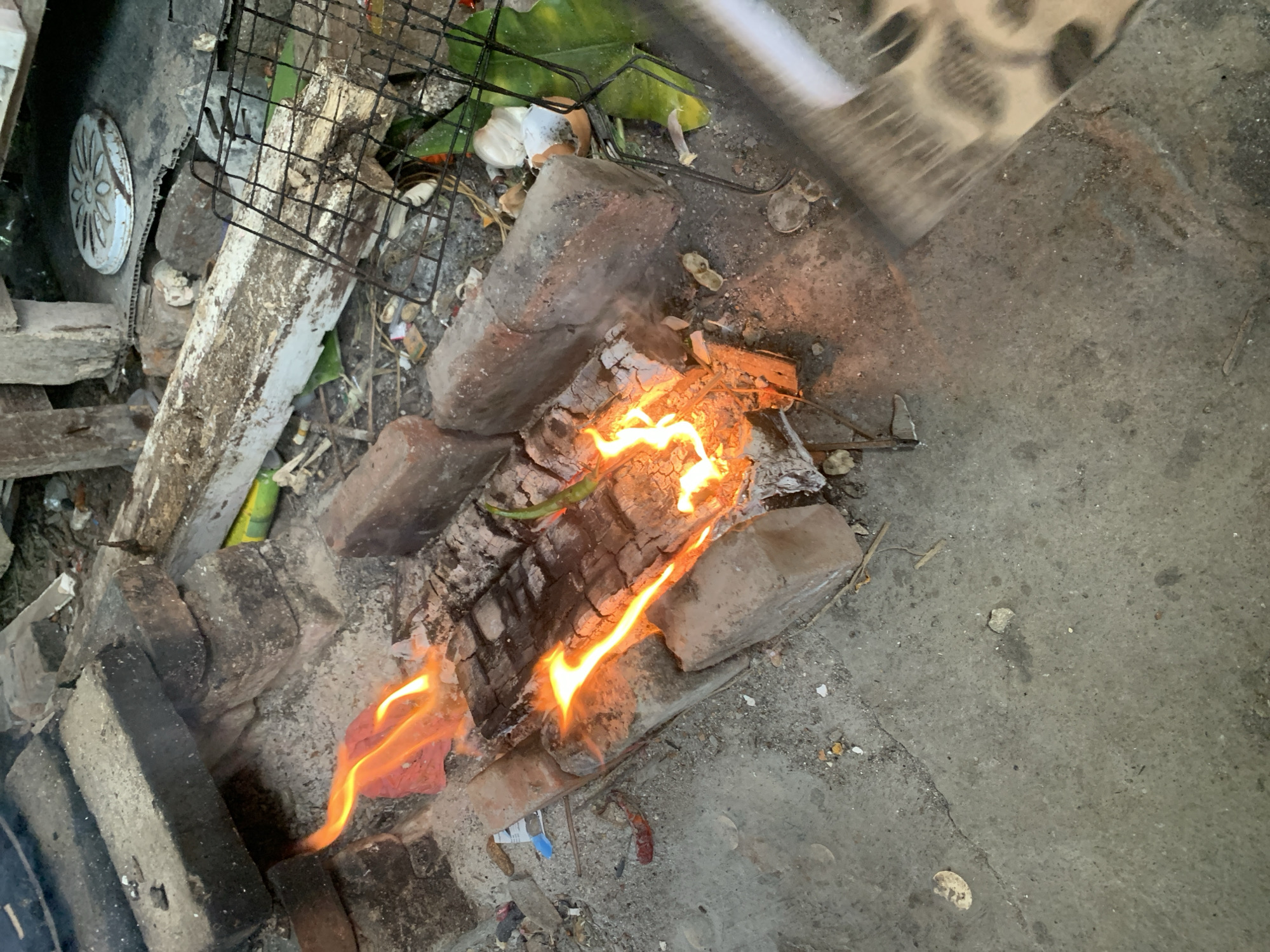 The image shows a small fire burning in a rudimentary hearth constructed from bricks and stones on a concrete surface. The location appears to be outdoors, with a grimy, possibly urban environment. A metal grill sits nearby, seemingly for cooking. A green leaf and what appears to be garlic are placed on the grill. The setting is in Karangkemiri, Indonesia.