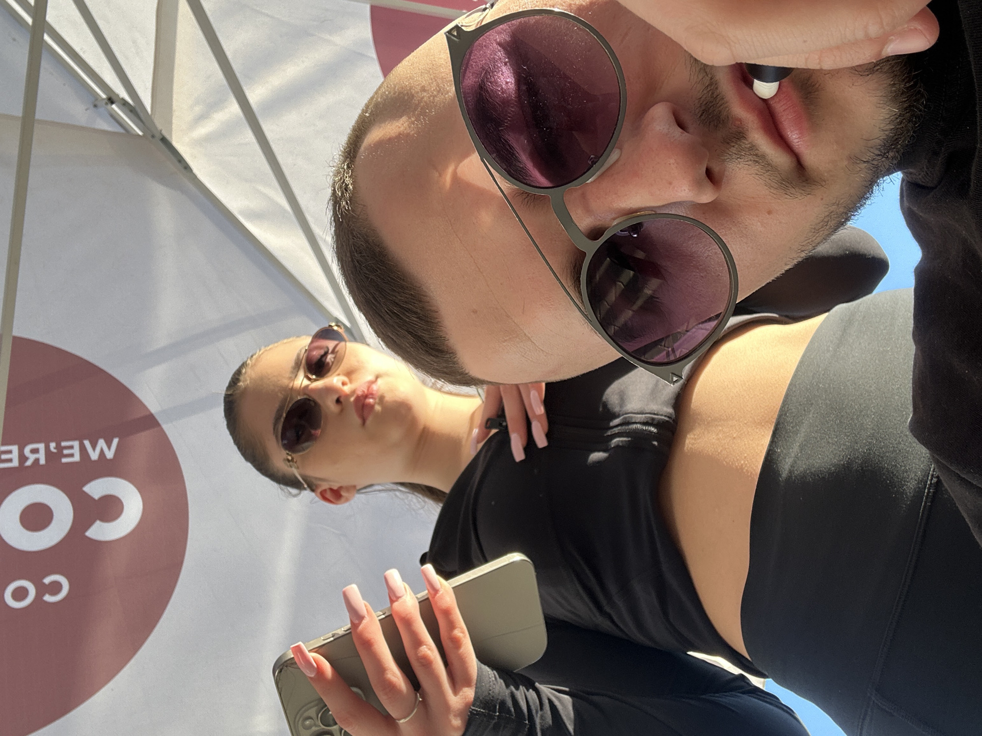 The image captures a low-angle selfie of two individuals outdoors under a white umbrella or canopy, suggesting bright daylight. In the immediate foreground, a person with short dark hair, some facial stubble or beard, and large round sunglasses with purple lenses looks down towards the camera. An earbud is held between their lips. They are wearing a dark top, possibly a hoodie.
Slightly behind and to the left, a woman with dark hair pulled back and wearing smaller, possibly aviator-style, sunglasses gazes towards the camera with a slight lip pucker. She is dressed in a black crop top, exposing her midriff, and long-sleeved athletic wear. Her hands, adorned with long, light-colored manicured nails and a simple ring on her left ring finger, hold a light-colored smartphone with a visible multi-lens camera array.
The large white umbrella above them shows its internal spokes and fabric. Reflected and reversed text is visible on the underside of the canopy, reading "WE'RE" and "CO CO," potentially part of a company logo like "WE'RE COOL" or "COFFEE CO." The background shows a clear blue sky. The scene conveys a relaxed, casual mood, likely depicting friends enjoying an outdoor moment in Plovdiv, Bulgaria.