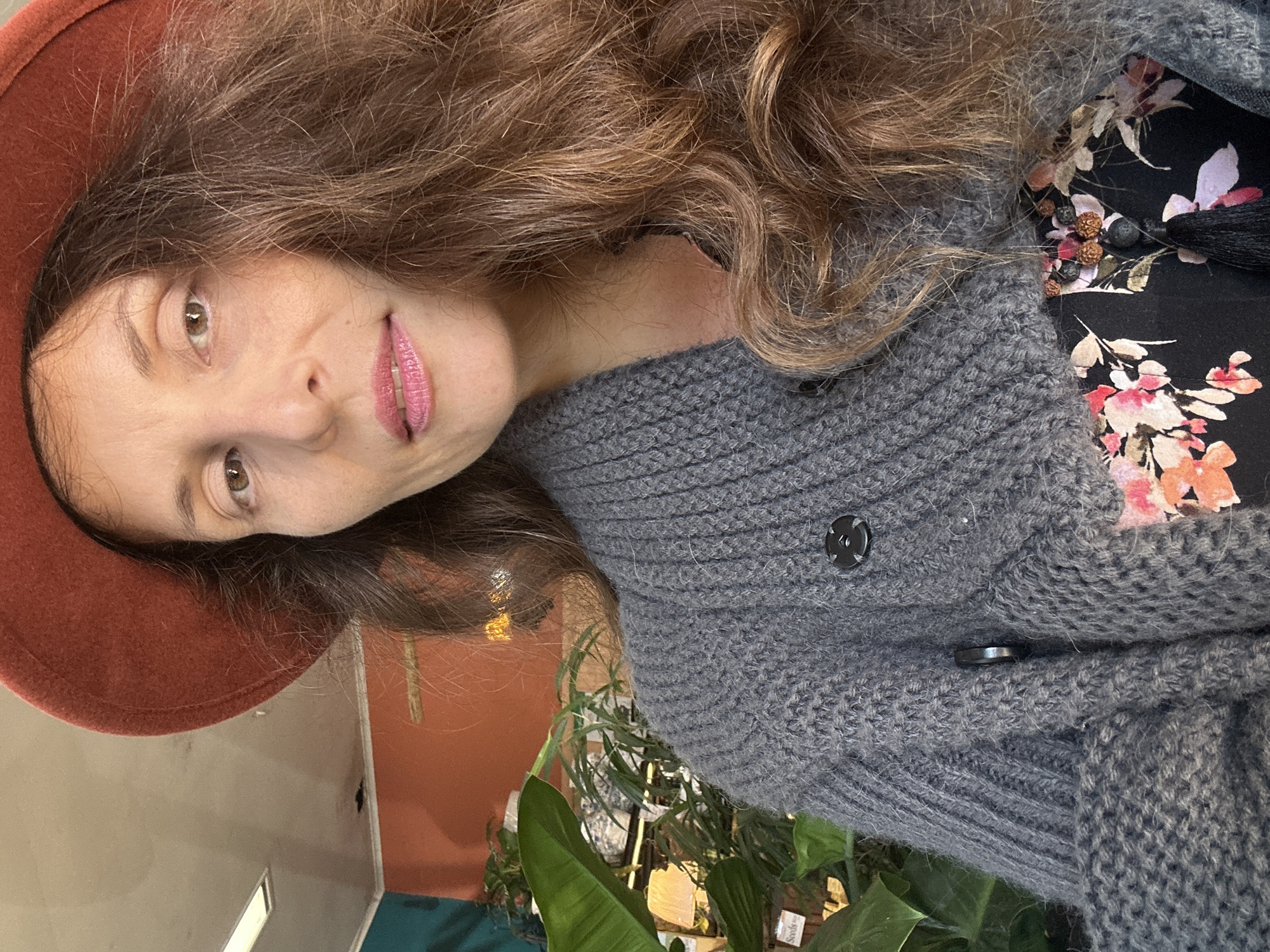 A woman wearing a rust-colored hat and a grey cardigan is pictured in a close-up shot. She has long, wavy brown hair and is looking towards the camera with her lips slightly parted. The background appears to be an indoor setting with plants visible. The location is specified as the city of Bellingen, Australia.