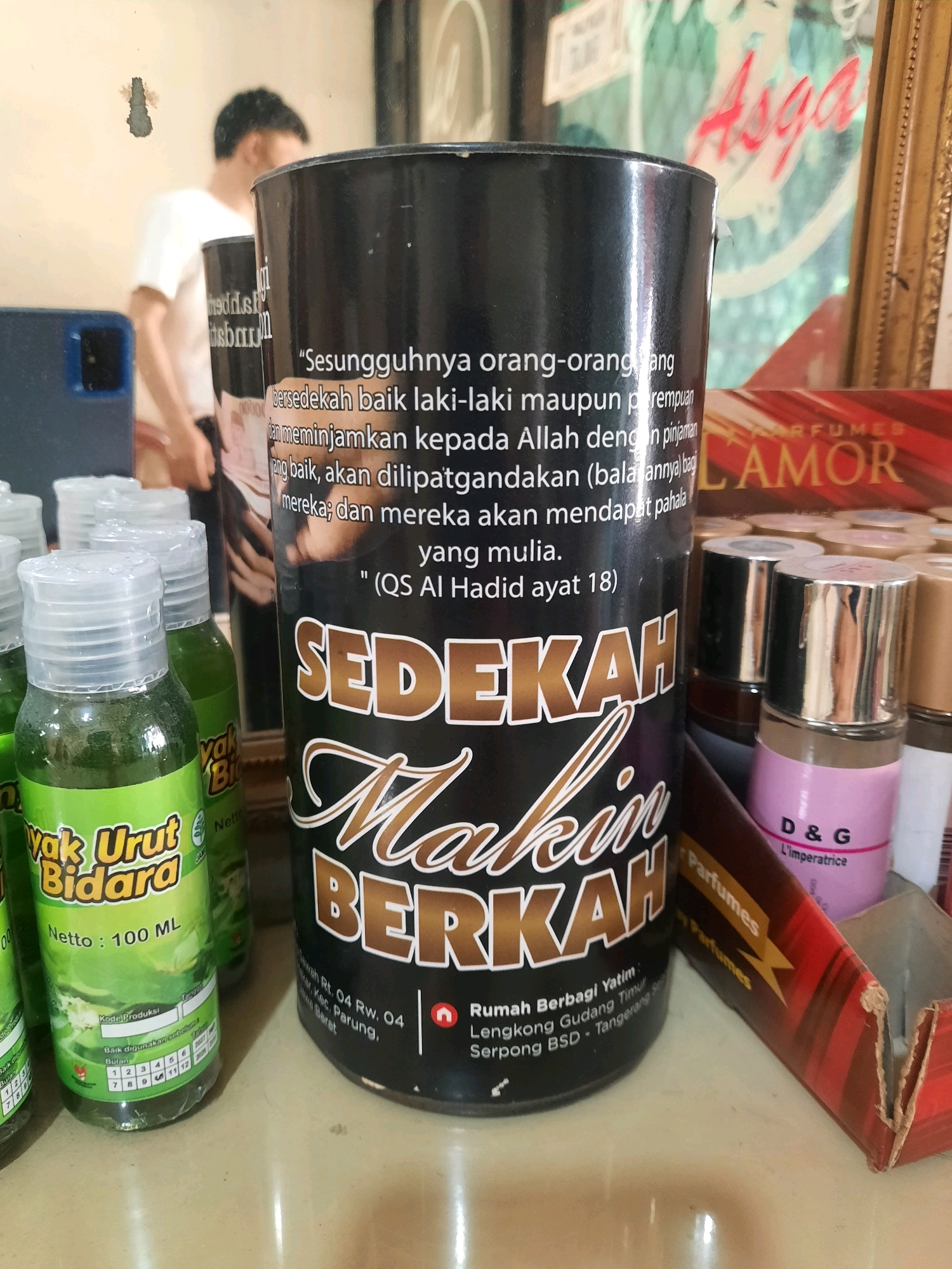 A close-up, eye-level medium shot shows a cylindrical black container with Arabic calligraphy on it, standing on a light-colored surface. The container is in the center of the frame, slightly to the right. The container has the text "SEDERAH MAKIN BERKAH" in large, gold, stylized font. Below this text, there is smaller text in white, which appears to be an address. The address reads "Jalan Rt. 04 Rw. 04, Rumah Berbagi Yatim, Lengkong Gudang Timur, Serpong BSD - Tangerang Selatan, Banten, Indonesia."
To the left of the container are several small green bottles with clear plastic caps. These bottles have green labels with yellow text. One visible label reads "YAK URUT BIDARA" and "Netto: 100 ML".
To the right of the container, there are several small perfume bottles with silver caps. One bottle is clearly visible, pink in color, with "D&G" and "L'Imperatrice" printed on it. Behind these perfume bottles, a red and gold box is partially visible, with "Parfumes" written on it.
In the background, a person wearing a white t-shirt is visible, partially blurred. There is also a framed picture on the wall behind the person, with red text that appears to say "Asga". The overall lighting suggests an indoor environment. The surface the objects are placed on appears to be a countertop or table.