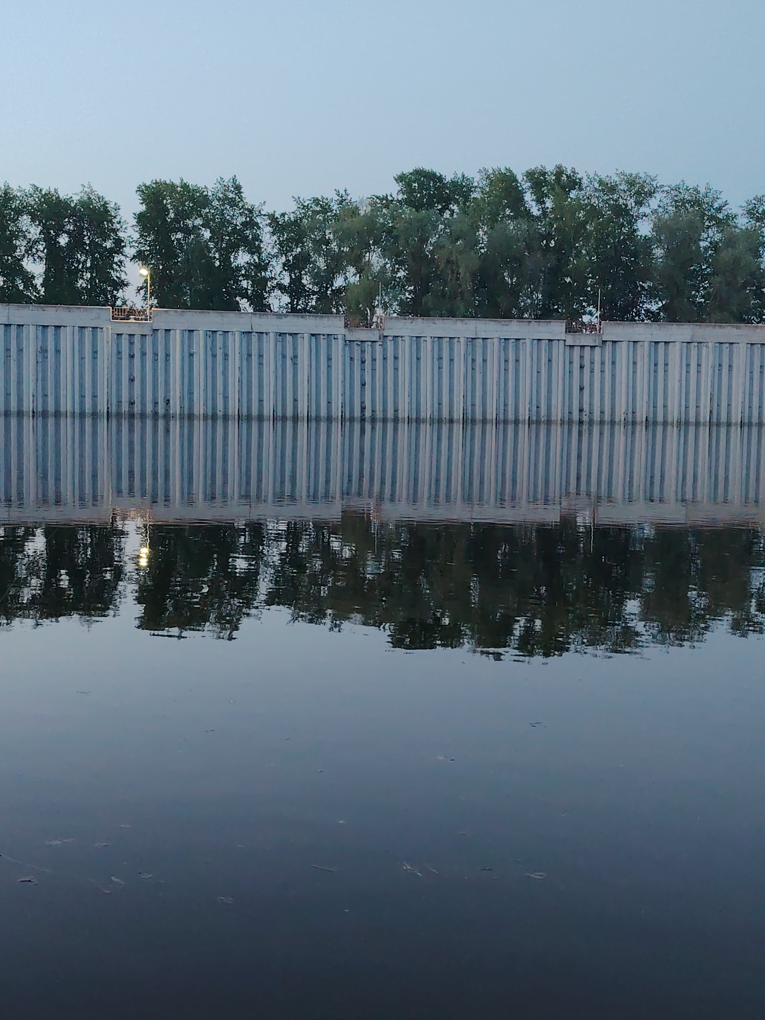 A still body of water reflects a concrete wall topped by a line of trees under a twilight sky. The wall is made of vertical panels with dark shadows between them. The reflection in the water is clear near the wall, but fades into a darker surface further away. The location is Chaikovsky, Russia.