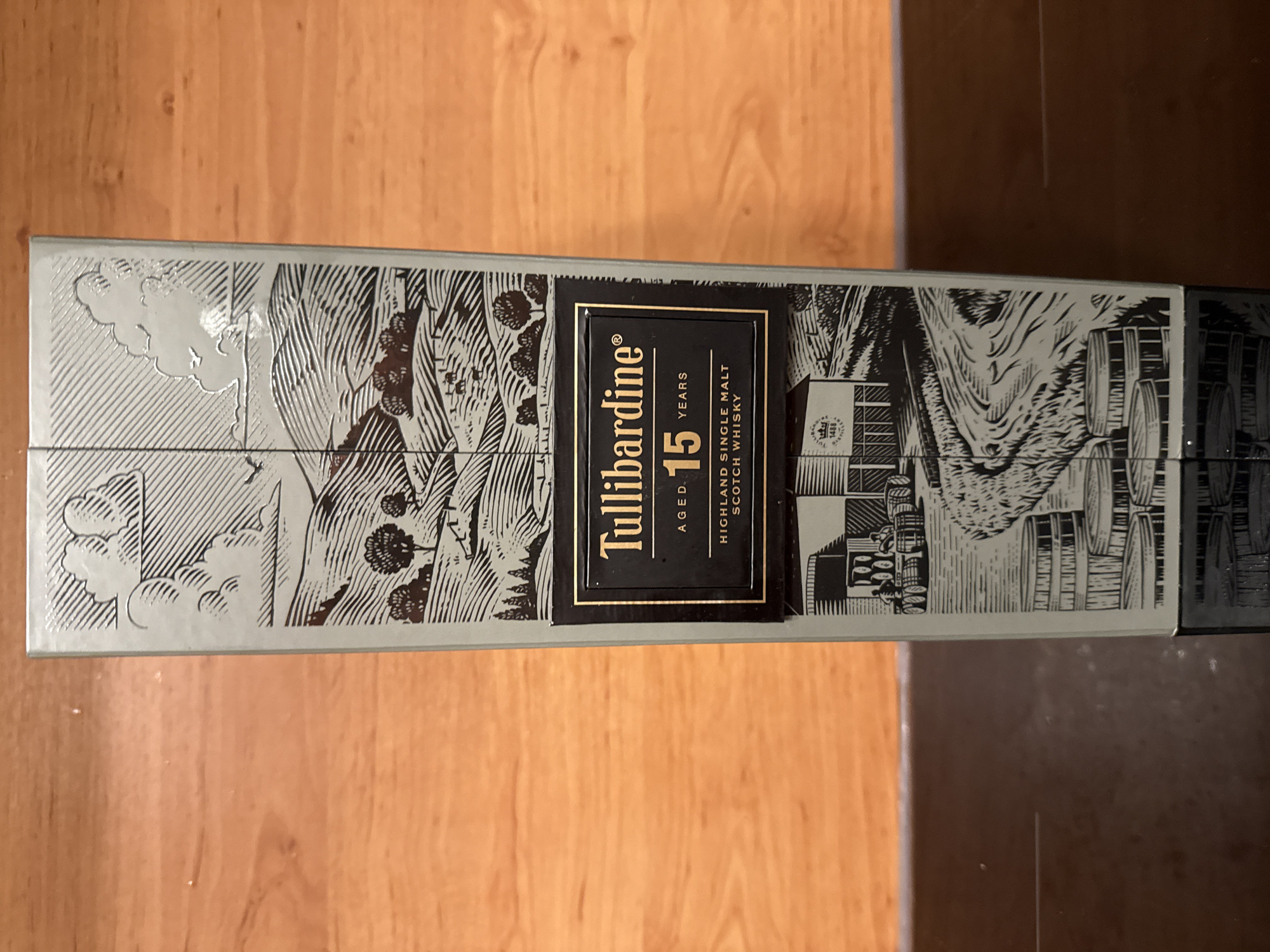 The image shows the side of a Tullibardine 15 Years Old Highland Single Malt Scotch Whisky box. The box has a detailed illustration of a rural landscape with rolling hills, trees, livestock, and a building that appears to be a distillery. There is also an illustration of barrels. The text on the box clearly states "Tullibardine", "AGED 15 YEARS", and "HIGHLAND SINGLE MALT SCOTCH WHISKY". The box is positioned vertically against a wooden background, likely a table or shelf. No people or any specific location in Silvolde, Netherlands are visible.