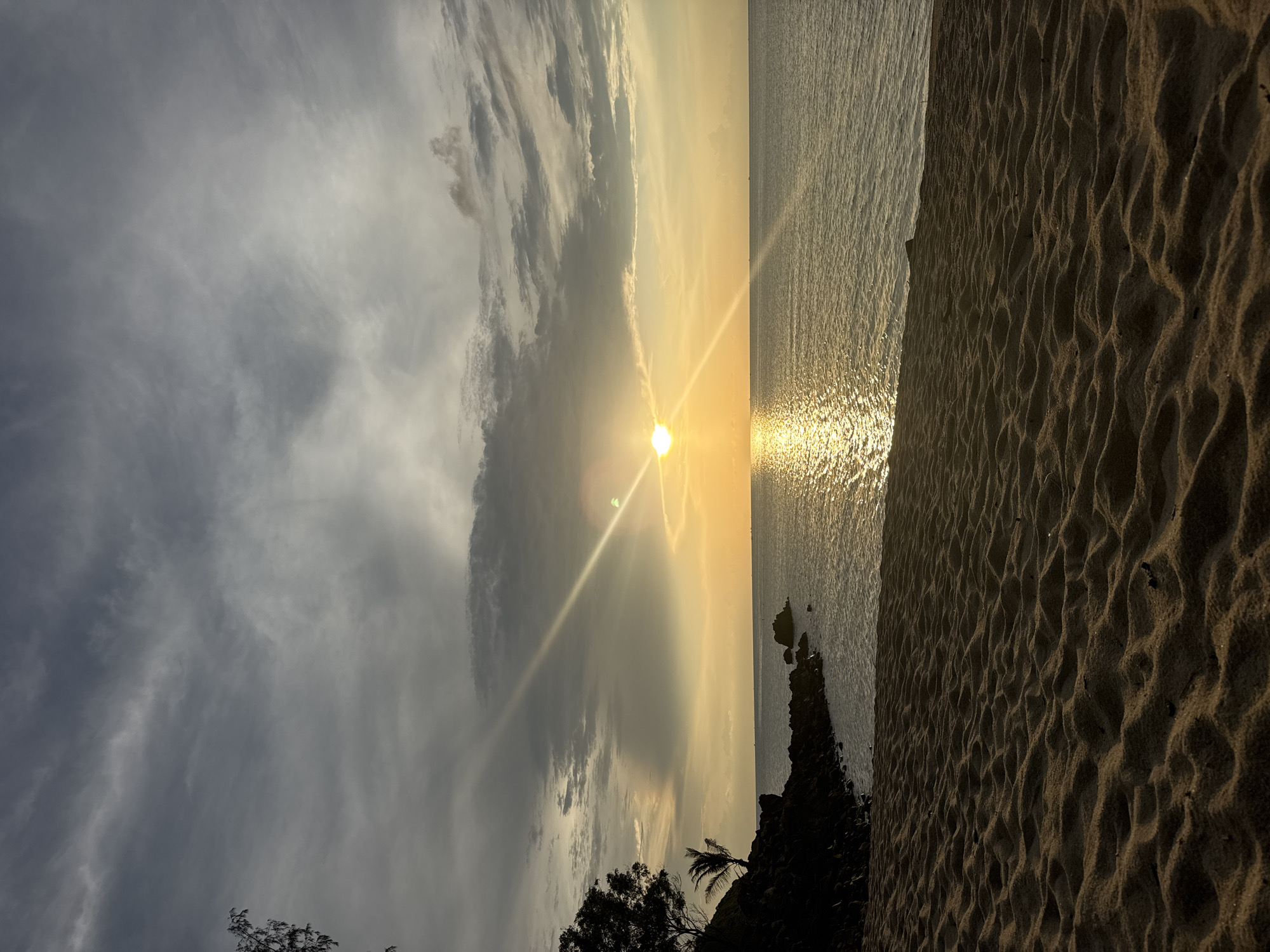 The image depicts a coastal scene in Baan Wang Ta Kien, Thailand, at sunset. The sun is positioned near the horizon, emitting bright rays that reflect off the water. The sky features a mix of clouds, some dark and others lighter, indicative of variable weather conditions. The water appears calm with visible sun glint. The foreground includes a sandy beach with textured patterns, and vegetation (trees, possibly palms) along the coastline. The orientation is likely vertical.