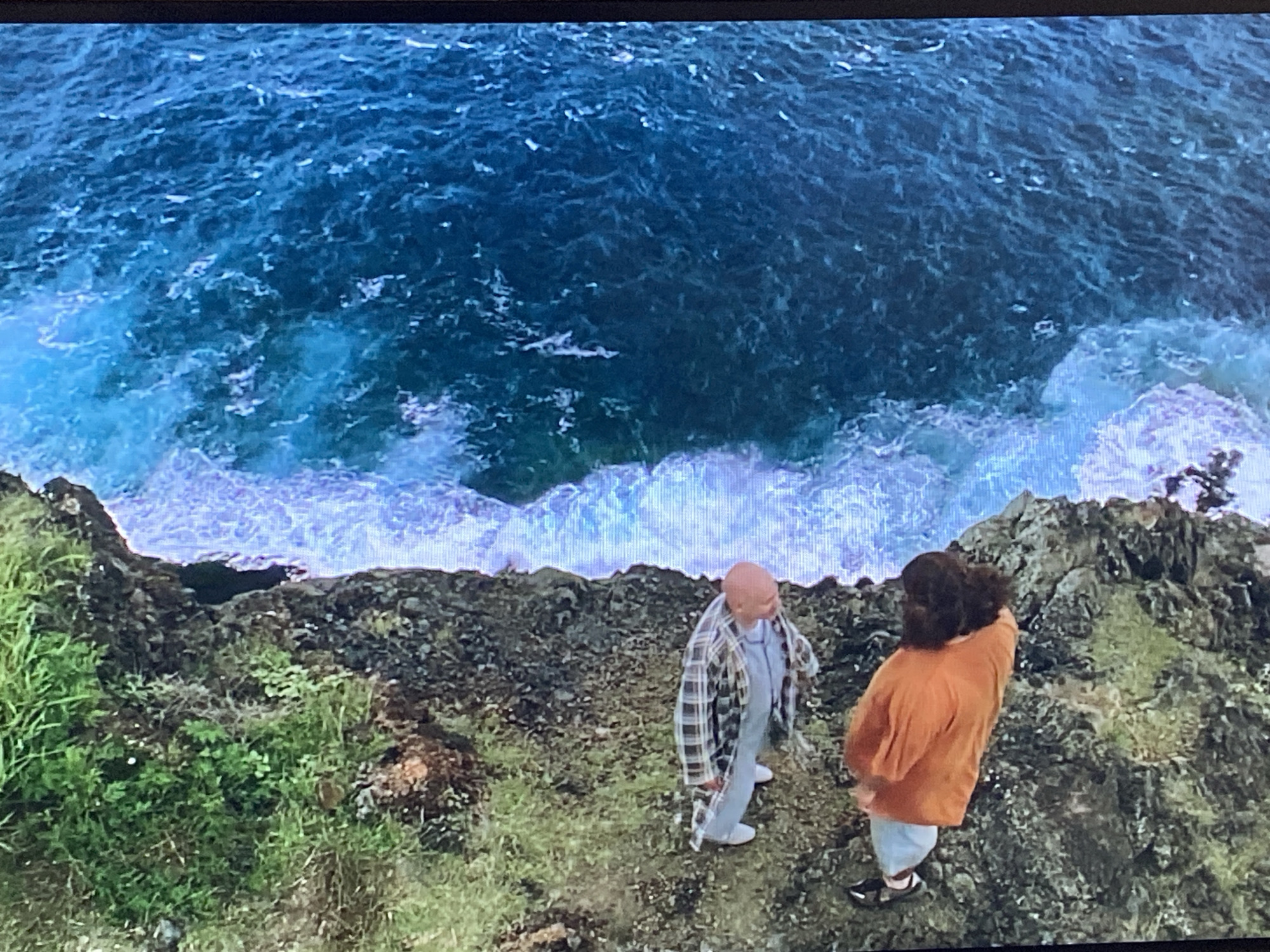 The image presents a high-angle view of two people standing on a rugged, grassy cliff overlooking a vast, turbulent blue ocean. The cliff face is rocky, interspersed with patches of green grass and sparse vegetation.
On the left, a bald individual wears a light-colored plaid shirt over a lighter inner shirt and light-colored pants, with white shoes. They appear to be looking towards the other person or out to sea. On the right, a person with dark, wavy hair wears an orange long-sleeved top and light-colored bottoms with dark footwear. This individual is turned away from the camera, gazing intently at the ocean.
The water below the cliff is a deep, rich blue, covered with numerous whitecaps and large, frothy waves that crash vigorously against the hidden rocks or shoreline, creating a dramatic white foam. The scene unfolds under what appears to be bright daylight. The primary activity is the two individuals observing the powerful and expansive ocean from their elevated viewpoint. There are no visible objects other than their clothing, and no discernible text in the image. The setting conveys a sense of wild, natural beauty and the raw force of the sea, consistent with a dramatic coastal landscape near Saint Petersburg, Russia.