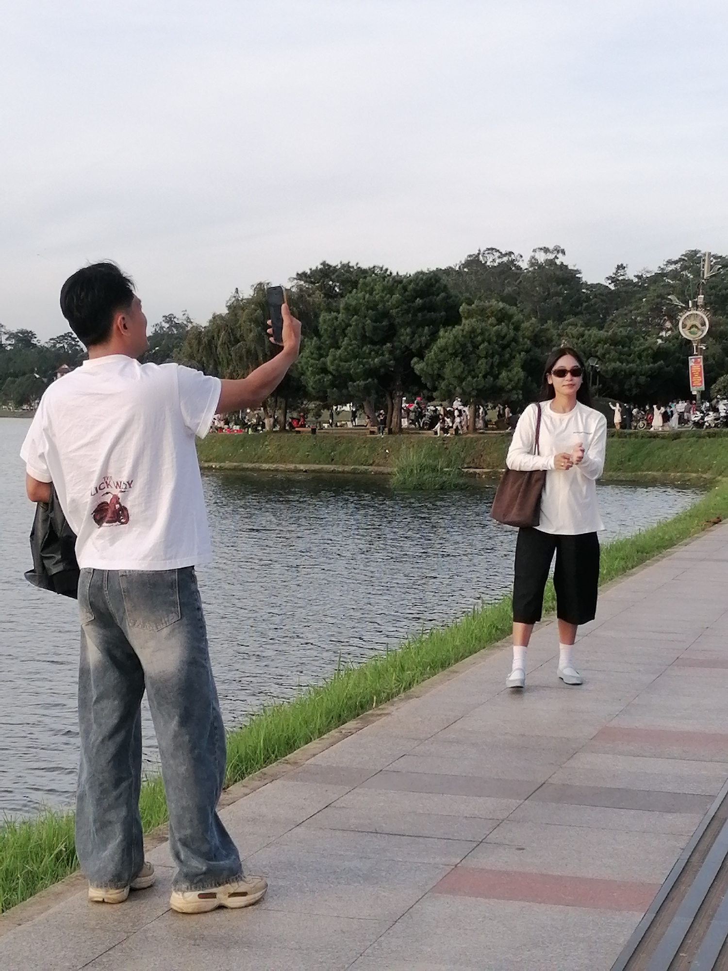 A man in a white t-shirt and loose-fitting jeans is taking a selfie with a smartphone in his right hand, while holding a black bag in his left. He is standing on a paved walkway next to a body of water. Further down the walkway, a woman in a white long-sleeved shirt, black culottes, and white socks is posing with her hands clasped in front of her. She is wearing sunglasses and carrying a brown tote bag over her left shoulder. In the background, a line of trees and a distant crowd of people are visible. The overall scene suggests a public park or recreational area, possibly Xuan Huong Lake in Da Lat, Vietnam, given the distinctive architecture of the lampposts and the lush greenery. The sky is overcast, indicating a cloudy day.