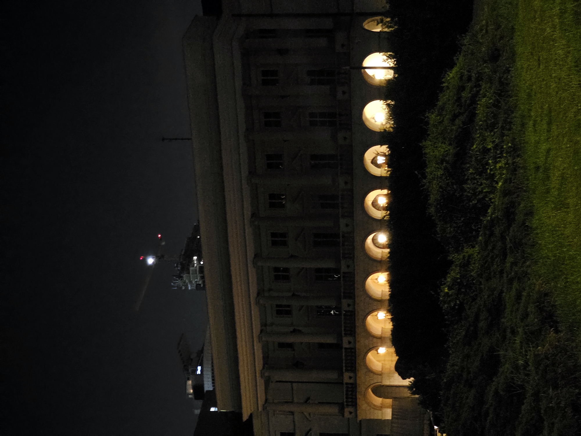 The image shows a building at night, likely in Warsaw, Poland. The building is a large, classical-style structure with columns and arched entrances at ground level. Several lights are visible within the arched openings, illuminating the scene. A dark, dense hedge runs along the front of the building, obscuring the lower portion of the structure. Beyond the hedge, a grassy area is visible in the foreground, also lit by the ambient light. In the background, the dark night sky is punctuated by a few distant lights and a construction crane. The overall impression is a nighttime view of an imposing, historic-looking building.
