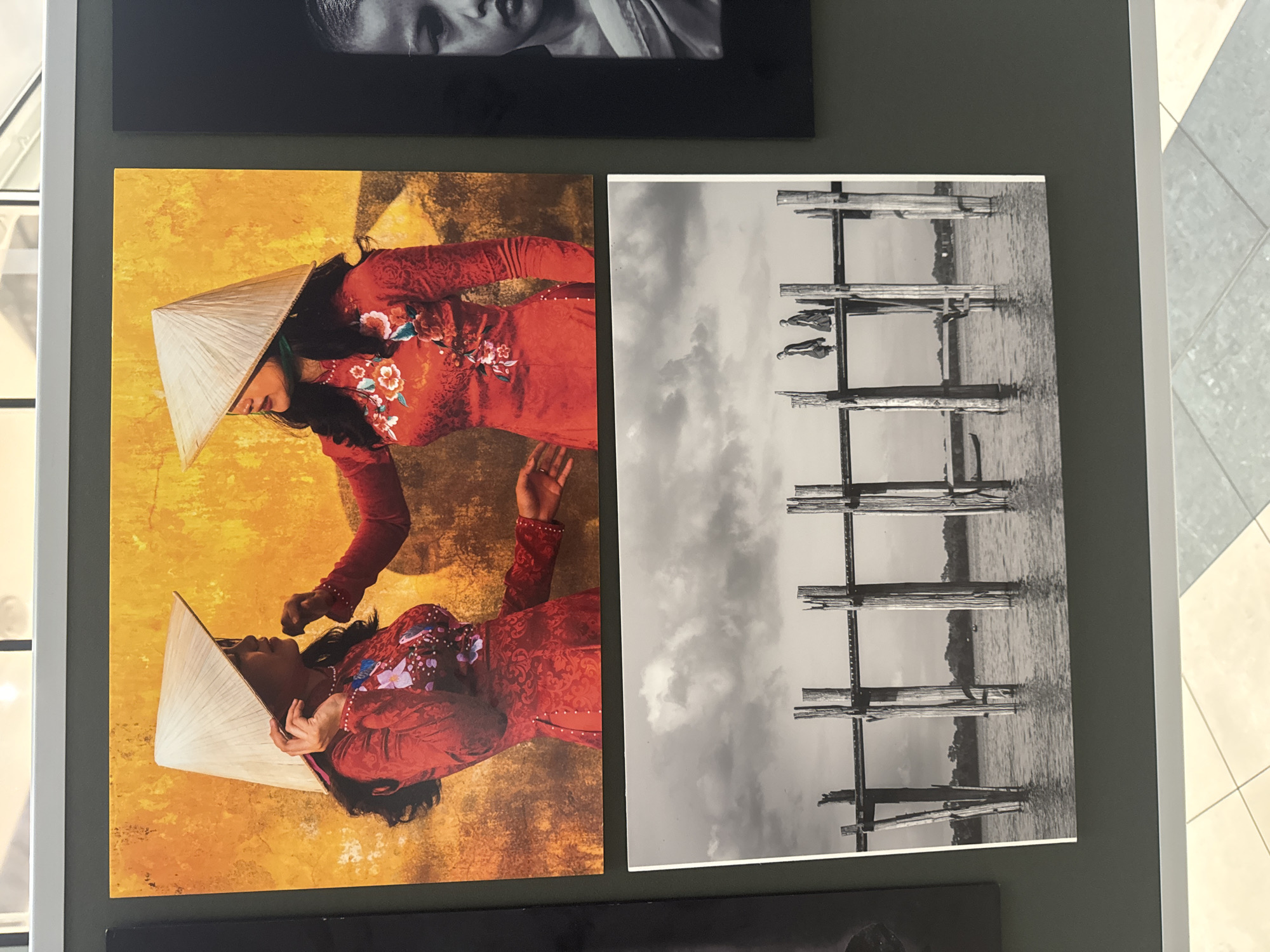 The image displays three photographs mounted on a dark grey wall, with a white frame at the top and bottom. The photographs are arranged in a vertical stack on the left and a single photograph on the right. The top left photograph is a colorful image featuring two women wearing traditional conical hats and red outfits adorned with floral patterns, standing against a textured yellow wall. The woman on the left is adjusting her hat, while the woman on the right reaches out towards her.
Below this, the bottom left photograph is a black and white image depicting a wooden bridge over water. Three figures, possibly monks in dark robes, are walking across the bridge, which is supported by numerous wooden posts. The water reflects the sky and the structure of the bridge. The right photograph is a black and white close-up of a young person's face, with dark hair and a serene expression.
The context provided indicates that this display is located in the city of Kilkenny, Ireland.