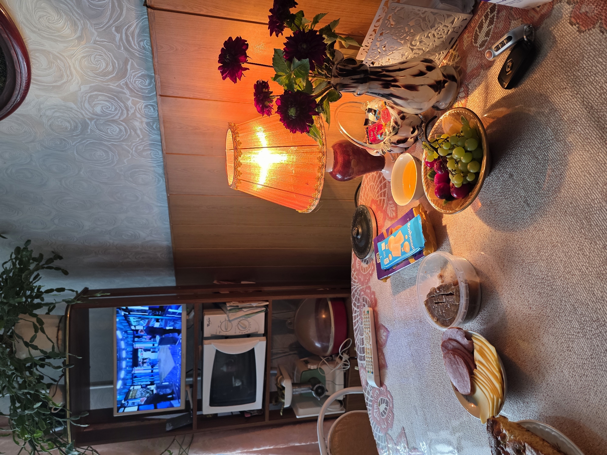 The image depicts a close-up view of a table laden with food and various objects, illuminated by a warm lamp. On the table, a slice of pie, a plate of sliced cheese and ham, a container of meat, a bowl of grapes and plums, and a small bowl of orange liquid are visible. To the right, a vase with dark flowers, a basket of candy, a decorative white holder, and car keys are present. A book or game box in blue and yellow is also on the table. In the background, a bookshelf holds a microwave and a television displaying a news program. The room has wallpaper with a rose pattern and wood paneling. The overall impression is of a domestic setting, possibly a dining or living area, with items suggesting a meal or snack is being enjoyed.