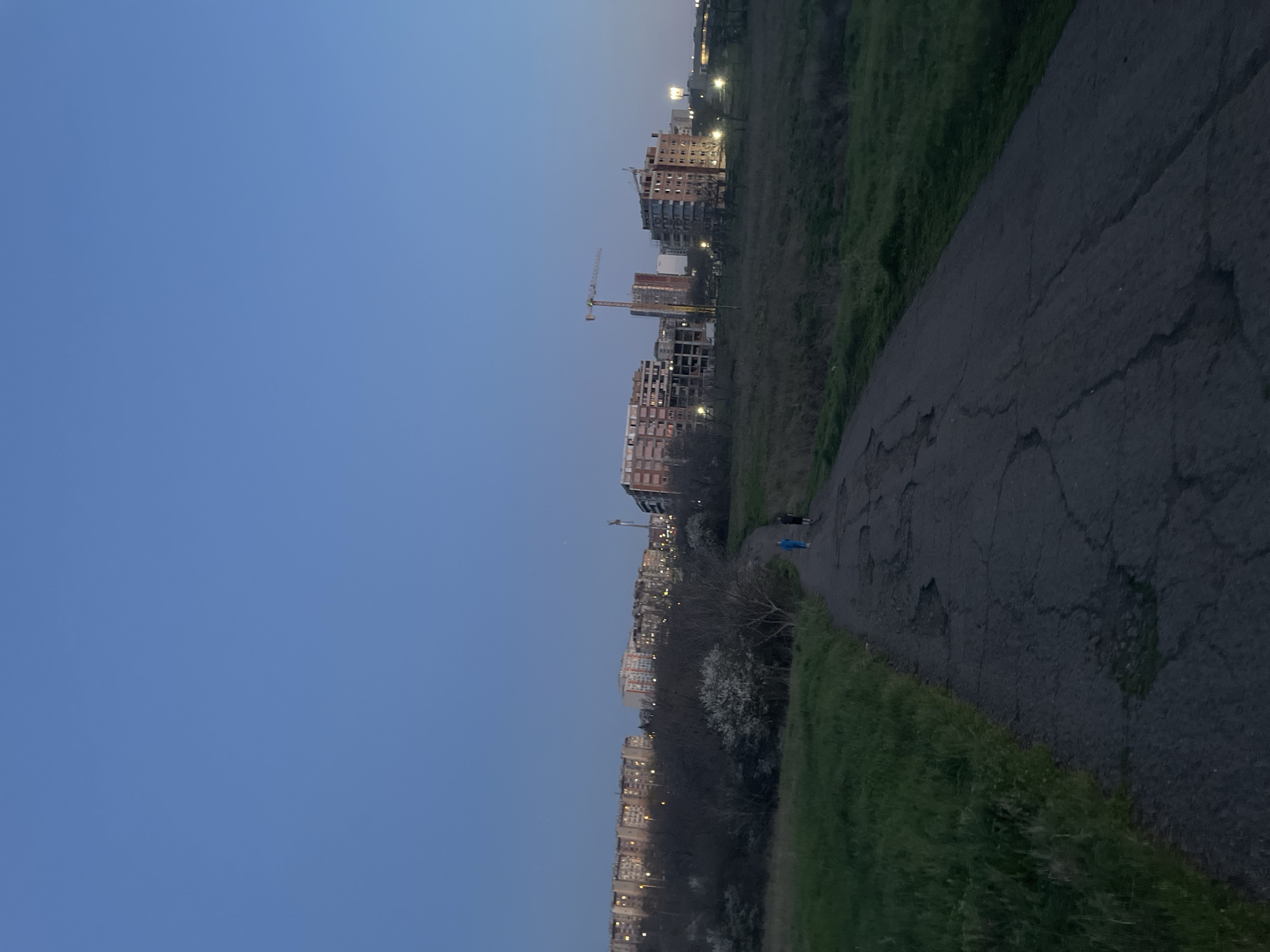 The image depicts a walkway in Burgas, Bulgaria, at dusk. The sky is a gradient of blue. To the right of the walkway is a grassy embankment, and to the left is a cityscape with several buildings, some under construction, indicated by cranes. Two people are visible on the walkway. The walkway appears to be asphalt and has cracks and patches. The environment is urban, bordering on natural.