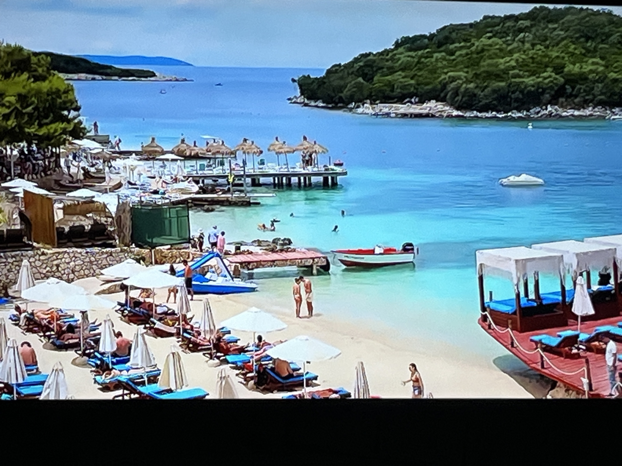 The image depicts a crowded beach scene with numerous sunbathers relaxing under white umbrellas on lounge chairs. The beach is a sandy expanse with clear, turquoise water. In the foreground, a red wooden structure with white canopies and blue seating is visible, with several lounge chairs on its deck. A few people are standing and walking on the beach, and some are in the shallow water. A red and white motorboat is docked near a wooden pier in the middle of the water. In the background, lush green trees cover a hillside that slopes down to the water. The sky is a pale blue. The scene is a tropical or Mediterranean resort area. The prompt asks to include the location in the city of Kharkiv, Ukraine, but the image itself does not provide any visual cues to suggest this location.