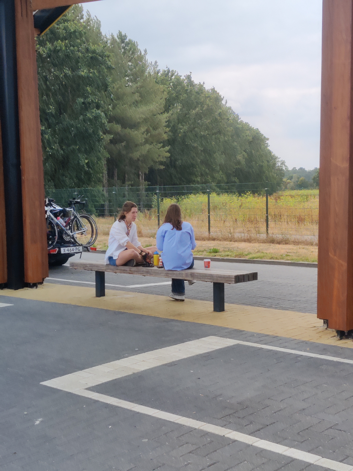 Two women are sitting on a wooden bench in an outdoor seating area next to a parking lot. The woman on the left is wearing a white shirt and blue shorts and is looking to her right. The woman on the right is wearing a blue shirt and dark pants and is looking away from the camera. A bicycle rack with two bicycles is attached to a black car on the left side of the image. The background consists of trees and a fence. The scene is located in the city of Maarheeze, Netherlands.