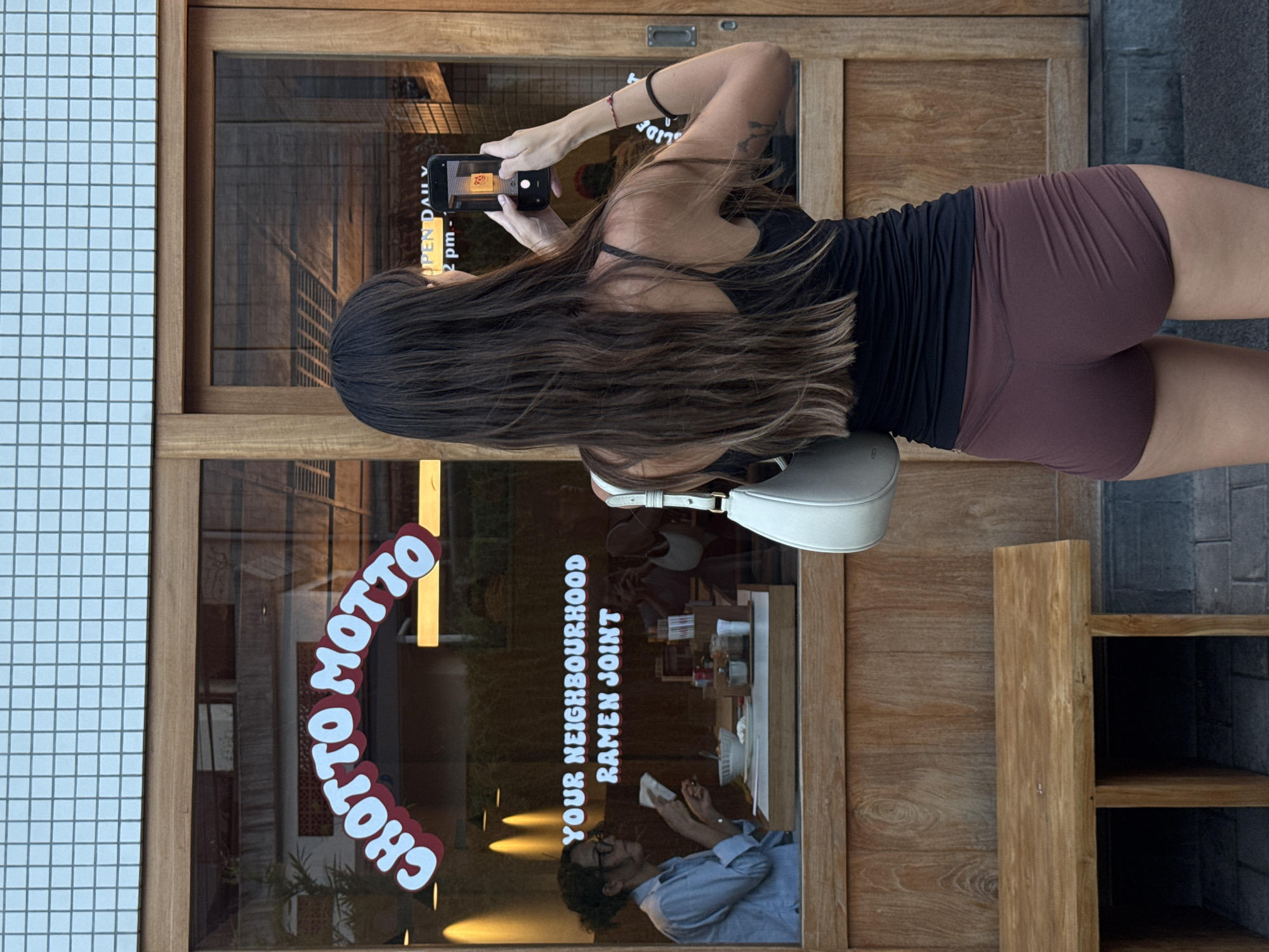 A woman is standing and taking a photo of "CHOTTO MOTTO YOUR NEIGHBOURHOOD RAMEN JOINT" restaurant signage and interior with her smartphone. The interior view shows a worker inside the restaurant. The photo was taken in Cemagi, Indonesia. The woman has long hair and is wearing a black tank top and brown shorts. She has a white purse on her shoulder.