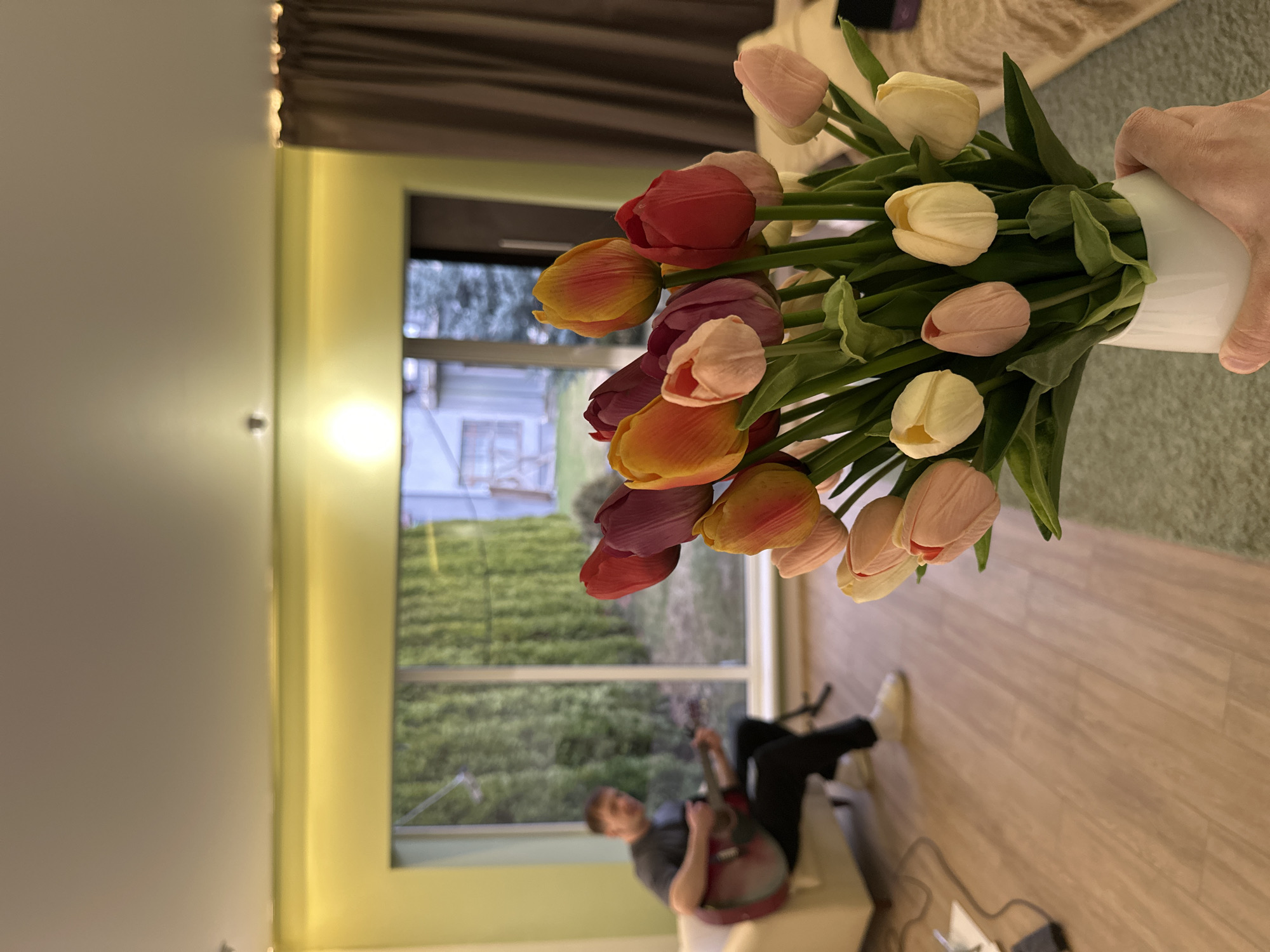 A person holds a vase filled with a bouquet of tulips in shades of red, orange, purple, pink, and yellow. A man sits in the background playing an acoustic guitar. The location is not possible to determine from this image.