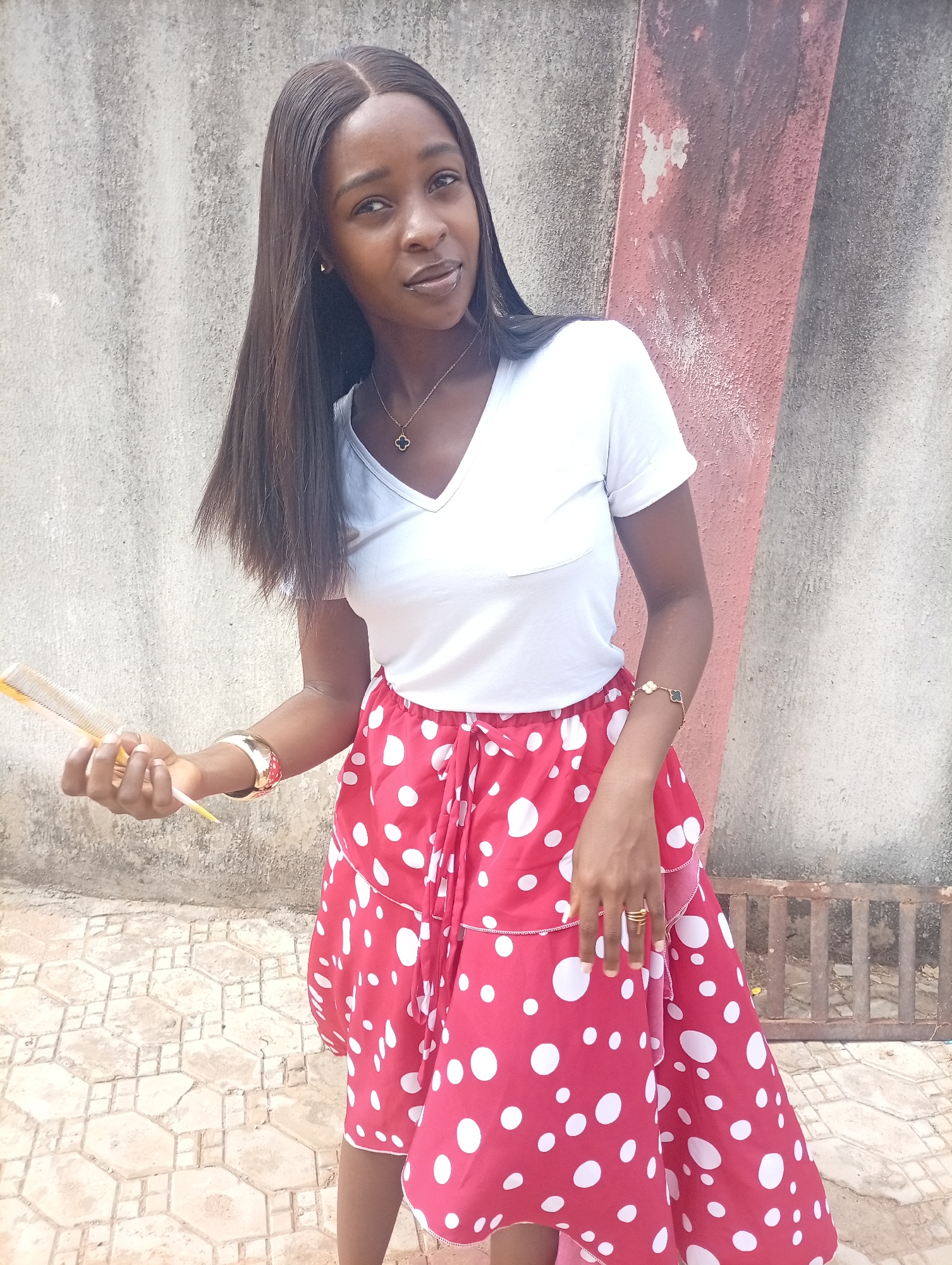 A young woman with dark hair and skin stands outdoors in what appears to be Rido, Nigeria. She is wearing a white V-neck T-shirt and a red skirt with white polka dots, tied at the waist. She holds a yellow comb in her left hand. The background includes a grey concrete wall with a reddish-brown vertical element to its right. A patterned tiled surface is visible underfoot. The subject wears a gold necklace with a black and gold pendant, a gold bracelet on her left wrist, a beaded bracelet on her right wrist, and gold rings on her fingers.