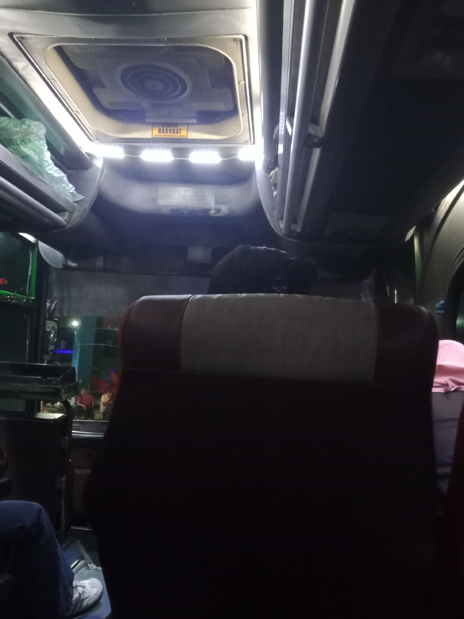 The image shows the interior of a bus, likely in Surakarta, Indonesia, as indicated by the text on a yellow sticker that reads "DARURAT" and "www.blemans.com". The bus appears to be in motion or parked at night, with the interior lights on. In the foreground, the back of a bus seat is visible, upholstered in dark brown and a lighter beige fabric. To the left, a person's legs are visible, wearing blue jeans and white sneakers. Further back, other passengers can be seen sitting in the bus. The ceiling of the bus features a ventilation grate and overhead compartments. The overall lighting suggests a dimly lit environment.