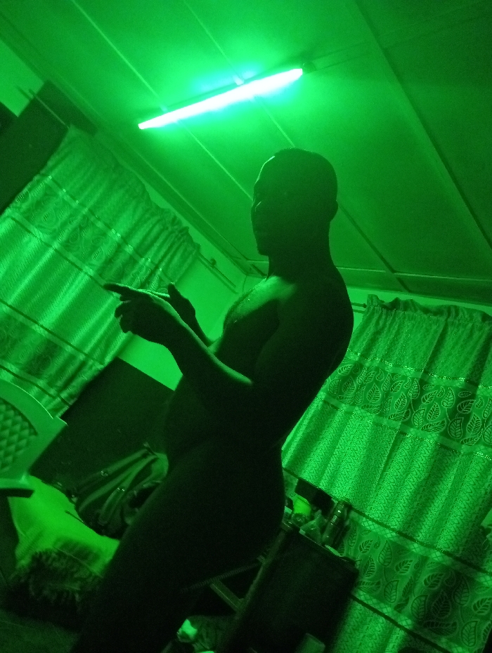 A person stands silhouetted against green-lit interior. The individual is nude from the waist up, pointing with their right hand. Curtains adorn a wall in the background. A fluorescent light fixture illuminates the ceiling. Furniture is visible but indistinct. The intense green lighting obscures details.