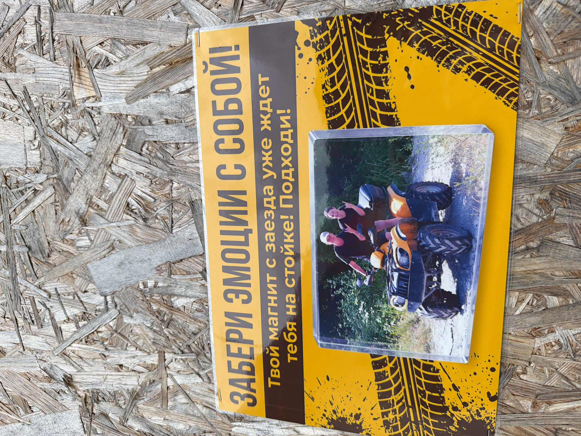 The image shows a yellow and brown rectangular promotional item or sign lying on a textured wooden surface. The top of the sign features large, bold black Cyrillic text that reads "ЗАБЕРИ ЭМОЦИИ С СОБОЙ!" (Take emotions with you!). Below this, in a smaller dark brown font, is the text "Твой магнит с заезда уже ждет тебя на стойке! Подходи!" (Your magnet from the ride is already waiting for you at the counter! Come over!). The lower portion of the sign is decorated with a yellow background featuring stylized black tire tracks and splatters. A clear, rectangular photo magnet is affixed to the sign, displaying an image of two people on an orange ATV in an outdoor, possibly wooded, setting. The person in the driver's seat, wearing a white cap and black t-shirt, is looking at the camera, while the passenger, in a dark t-shirt, is giving a hand gesture. The promotional item is designed to advertise a magnet related to an ATV ride. The location is stated to be the city of Shepsi, Russia.