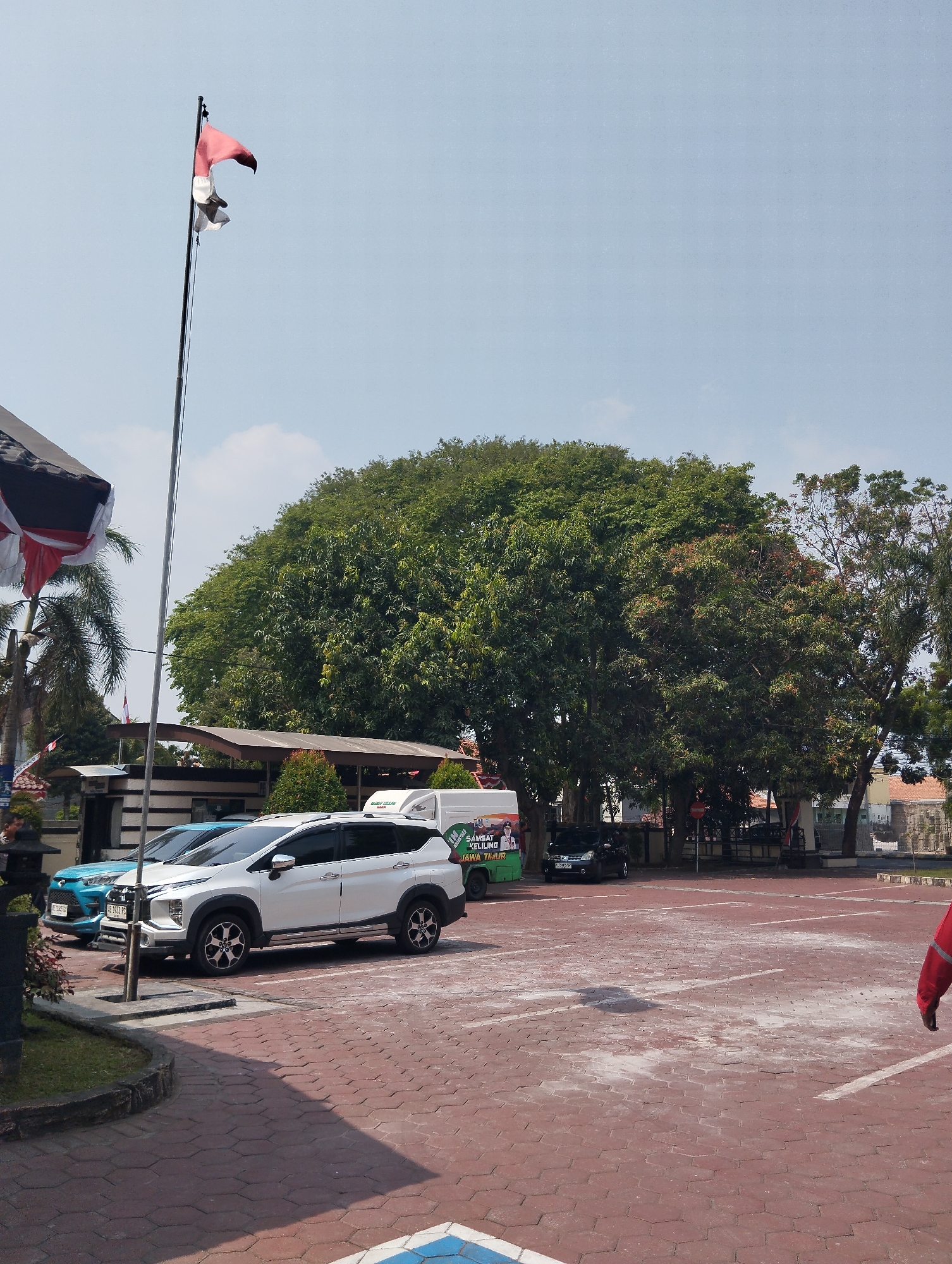 A mid-shot, eye-level, vertical outdoor photograph captures a parking lot in Madiun, Indonesia, under a clear blue sky. In the foreground, a textured red brick-patterned paving occupies the ground, marked with white parking lines. To the left, a grassy area with a concrete border is visible. Dominating the mid-ground are several parked vehicles, including a white SUV and a turquoise sedan. Behind the cars, a large, leafy green tree stands prominently. Further back, a low-slung building with a brown roof and striped walls is present, along with more trees and a glimpse of distant buildings. A tall flagpole with a red and white flag, the flag of Indonesia, stands to the left of the frame. The scene appears to be a public space, possibly an office or administrative building parking lot, given the presence of the Indonesian flag and the general layout.