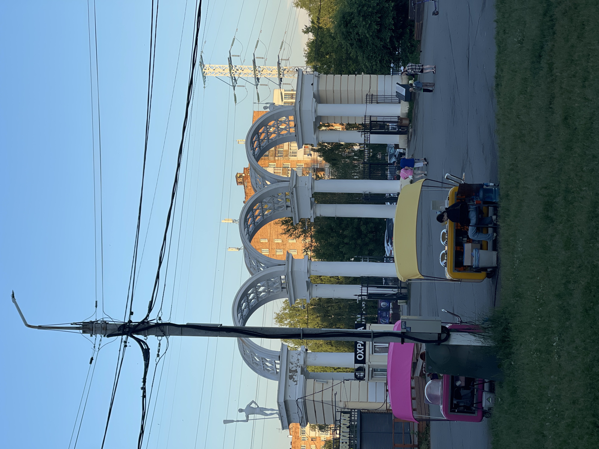 In an outdoor urban setting, a large arched gateway with multiple pillars and decorative arches stands prominently. A statue of a person holding a paddle is visible on top of the leftmost pillar. Two colorful food carts, one pink and one yellow, are parked in front of the gateway. The yellow cart has a person seated behind it. Several individuals are walking or standing near the gateway and in the background. Overhead, a complex network of electrical wires is supported by a tall pole and stretches across the sky, with a large electrical transmission tower visible in the distance. Residential buildings and lush green trees form the backdrop. The ground is paved with asphalt. The lighting suggests it is late afternoon or early evening.