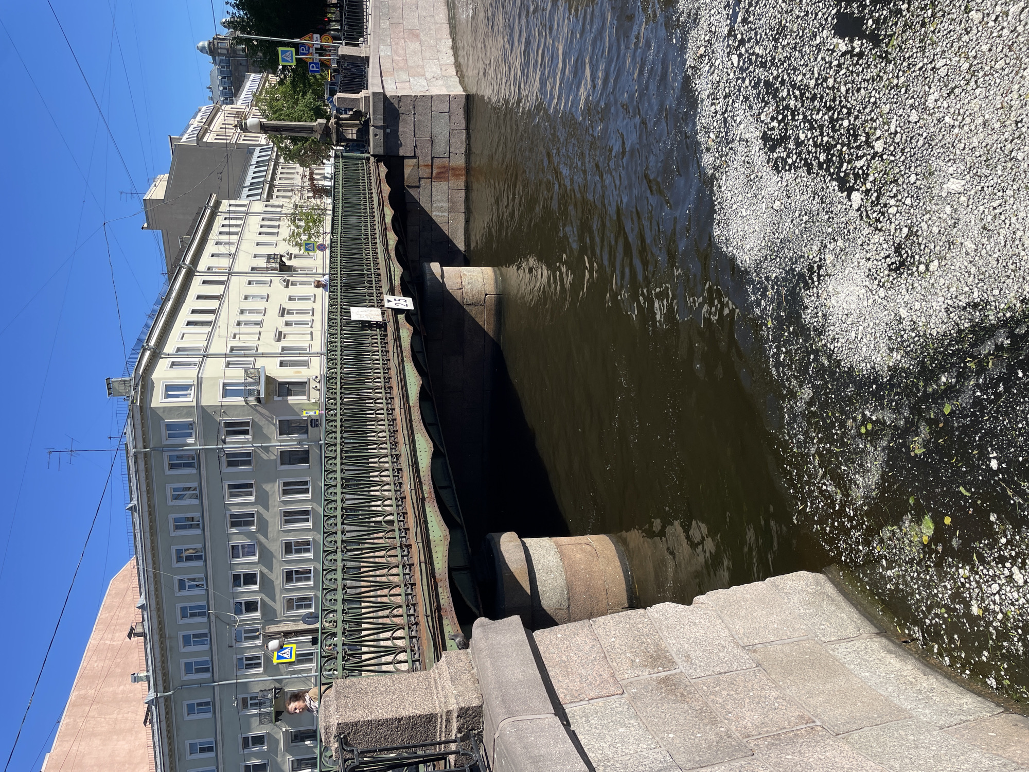 The image shows a view of a canal in Saint Petersburg, Russia, on a sunny day. In the foreground, there are stone bridge supports and paving stones. A green metal fence lines the edge of the canal.
To the left of the canal, there is a multi-story building with many windows, painted a pale yellow color. Across the canal, there is more architecture visible, including rooftops and domes, suggesting an urban setting typical of Saint Petersburg. A blue sky dominates the upper portion of the image, indicating clear weather.
The canal water appears dark and somewhat murky, with a significant amount of light-colored debris, possibly foam or natural material, floating on the surface towards the right side of the frame. The lighting suggests it is midday, with strong shadows cast by the bridge and buildings.
There are a few people visible. One person is standing on the bridge walkway, looking out towards the canal. Another person is partially visible near the base of the building on the left.
Several signs are visible. One sign on the building appears to have text and possibly a pictogram. Another yellow diamond-shaped sign with a pedestrian symbol and an arrow is mounted on a pole near the building. A small white sign with the number "25" on it is attached to the fence. The overall impression is a glimpse of everyday life in a historic European city.