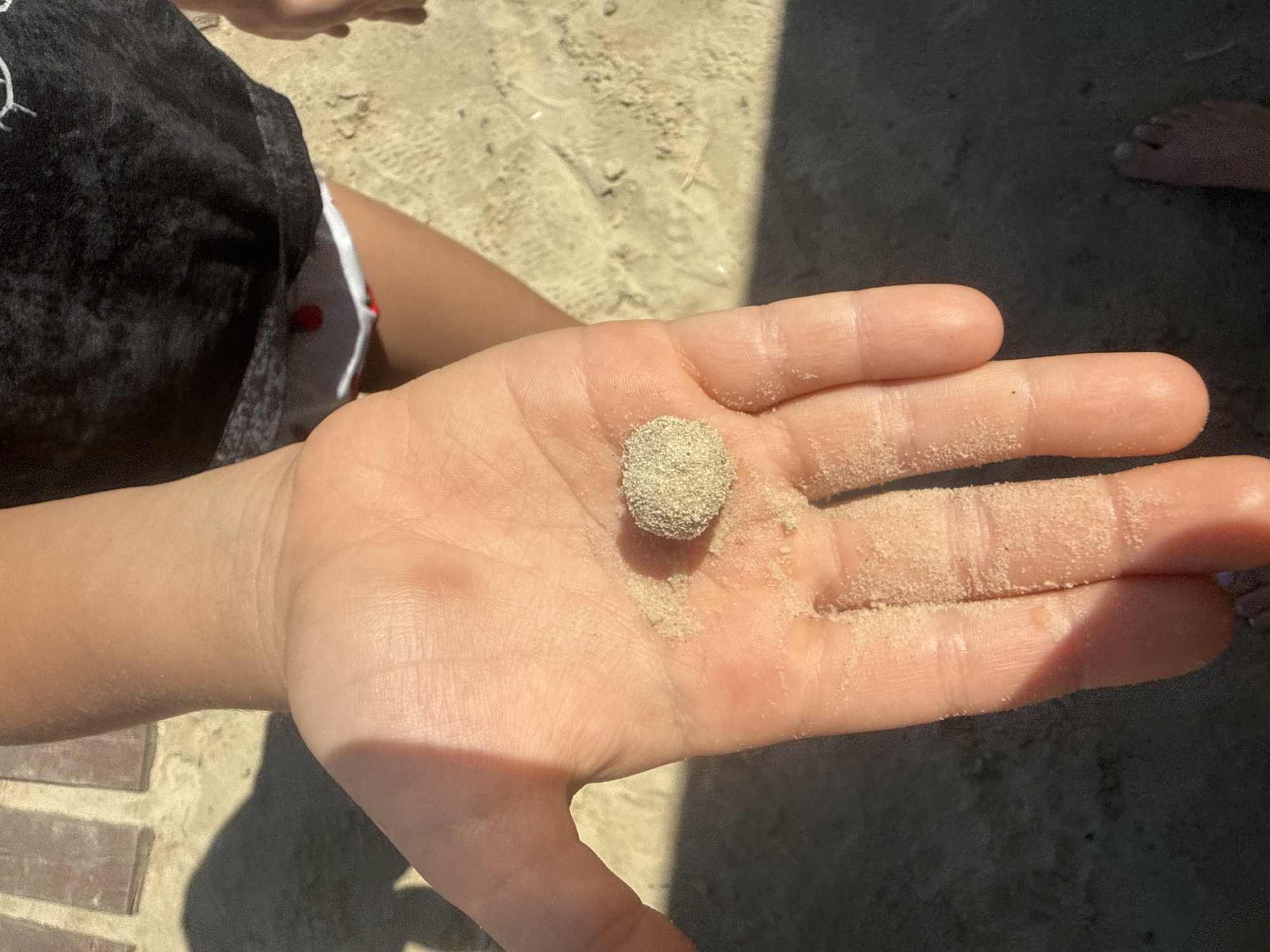 The image shows a close-up of a person's hand holding a small, spherical clump of sand. The hand is open, palm up, and is lightly dusted with sand. The sand ball is positioned in the center of the palm. The background is out of focus but appears to be a sandy surface, possibly a beach, with some shadows and what looks like a person's foot in the upper right corner. The lighting suggests it is daytime, with sunlight casting shadows. The overall tone of the image is casual and summery, evoking a sense of being at the beach. No text is visible in the image.