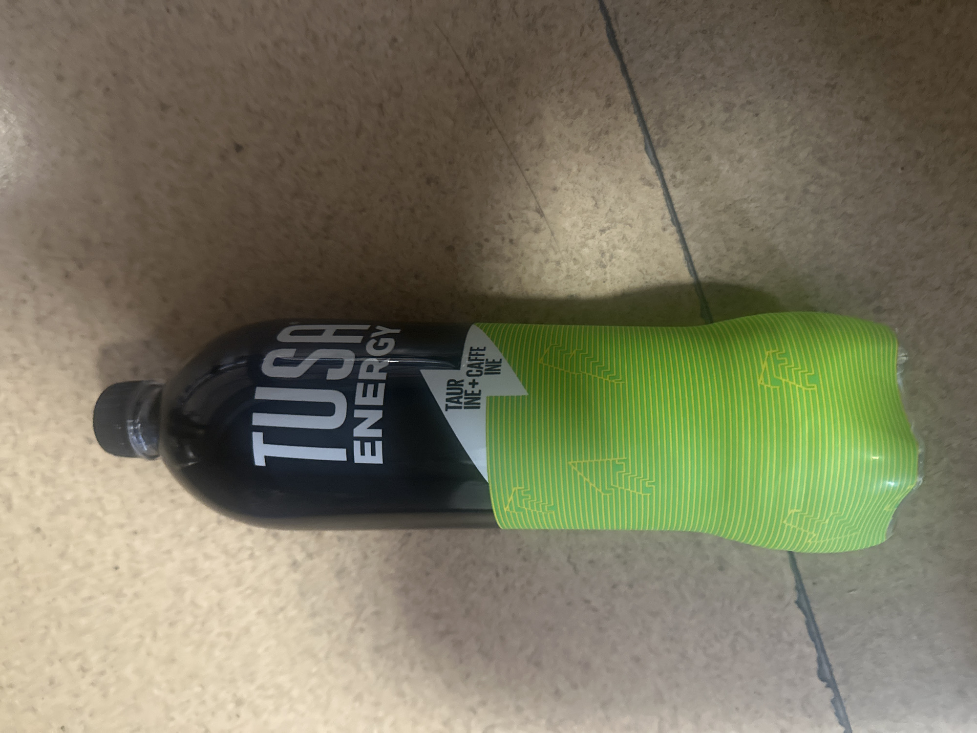 A tall, black plastic bottle of TUSA ENERGY drink with a bright green bottom section featuring a geometric pattern and images of lightning bolts. The bottle has a black cap. The text on the bottle reads "TUSA ENERGY" in large white letters, and below that, in smaller white text on a triangular white label, are the words "TAUR INE+CAFFE INE". The bottle is standing upright on a light brown, textured floor that appears to be made of concrete or a similar material, possibly in a residential or commercial setting in Novosibirsk, Russia. The lighting suggests an indoor environment. There are no people or animals present in the image.