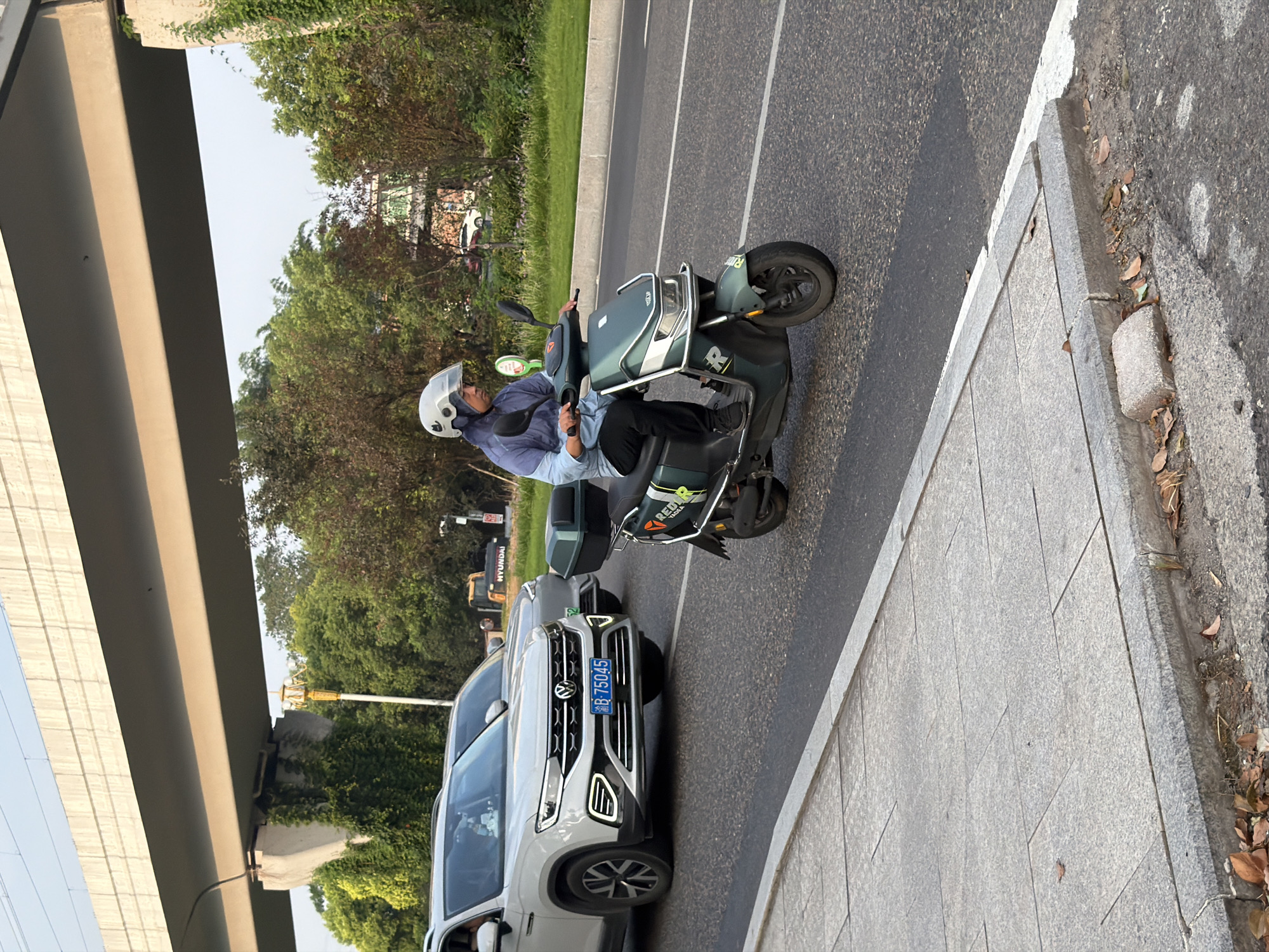 In the city of Jiangjin, China, a person wearing a light blue long-sleeved shirt, dark pants, and a white helmet rides a dark green electric scooter on a paved road. The scooter has a black storage box on the back and green and white decals on its side. In the background, a white SUV with a blue license plate displaying "B 75045" is parked. Further back, a grey SUV is also visible, along with a yellow excavator and trees. Overhead, a concrete overpass supported by pillars runs across the scene, with trees and vegetation growing on and around the pillars. The foreground shows a sidewalk made of grey paving stones next to the road, with fallen leaves scattered on the ground. The sky is clear and bright.