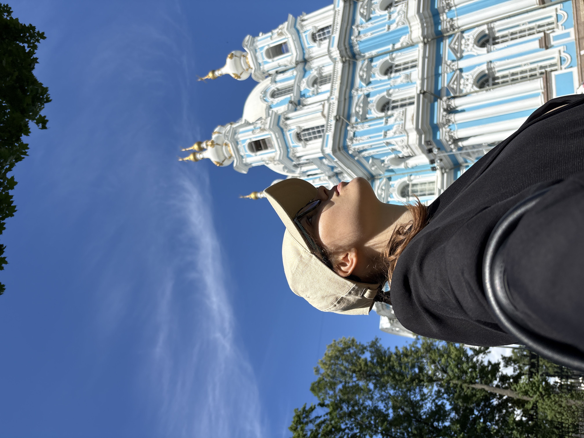 This is an eye-level, medium shot of a person looking up at the Smolny Cathedral in Saint Petersburg, Russia. The person is wearing a black jacket and a beige baseball cap. They are seen from the chest up, with their head tilted back and their gaze directed upwards. The sun is shining brightly, casting shadows and highlighting the intricate details of the cathedral's blue and white facade, which features ornate white detailing and gold accents on its multiple domes. The sky is a clear, vibrant blue with a few wispy white clouds. Green trees are visible in the background, framing the lower part of the image. The photo appears to be taken on a sunny day, likely in the late morning or early afternoon, given the strong sunlight. The person's upward gaze and the grand architecture of the cathedral suggest a sense of awe or admiration. No text is visible in the image.