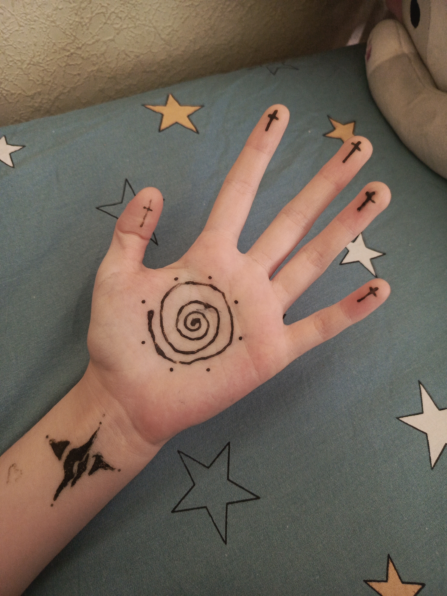 A person's hand is displayed on a star-patterned blue fabric. The person has tattoos. A star-shaped tattoo is visible on the wrist area of the arm. A spiral tattoo is located on the palm of the hand. Each finger has a cross tattoo near the fingertip. Part of a stuffed animal is present in the background. The setting is Unknown, Unknown.