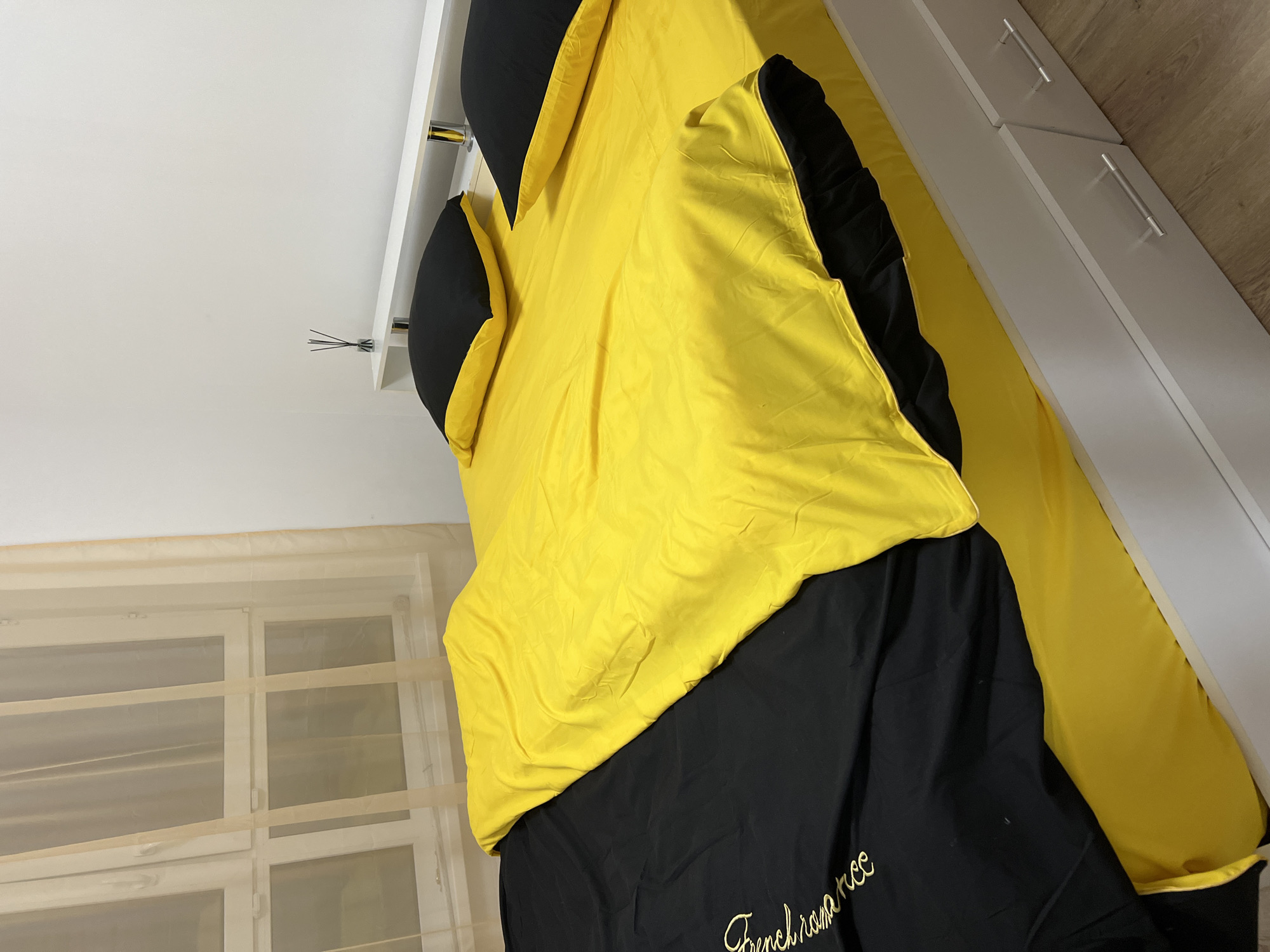 The image shows a bed with black and yellow bedding in a room. The bedding is folded over, revealing a black sheet with gold cursive text that appears to read "French romance." Two black pillows with yellow trim are visible on the bed. In the background, there is a white closet with silver handles and a window with sheer, beige curtains. The lighting suggests it could be daytime, and the presence of bedding indicates a bedroom setting. There are no people or animals visible in the image.