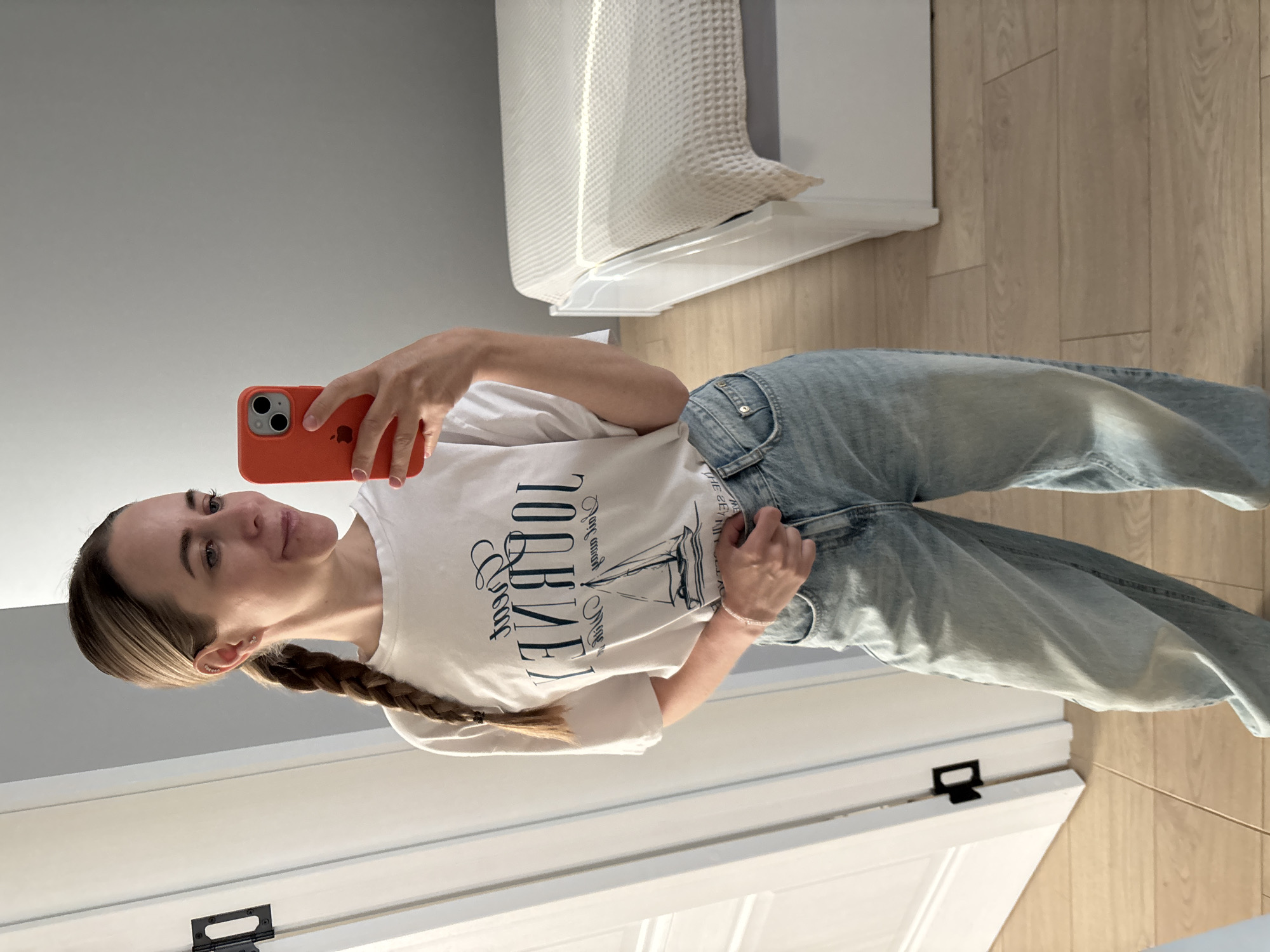 A young woman is taking a selfie indoors. She is holding an orange phone in her right hand. She is wearing a white t-shirt with a printed design and light blue jeans. Her hair is blonde and styled in a braid. The room appears to be a modern interior, with light wood flooring and a gray wall. In the background, there is a white piece of furniture, possibly a bed or storage unit, and a white door with black hardware. The photograph was likely taken in Chaikovsky, Russia.