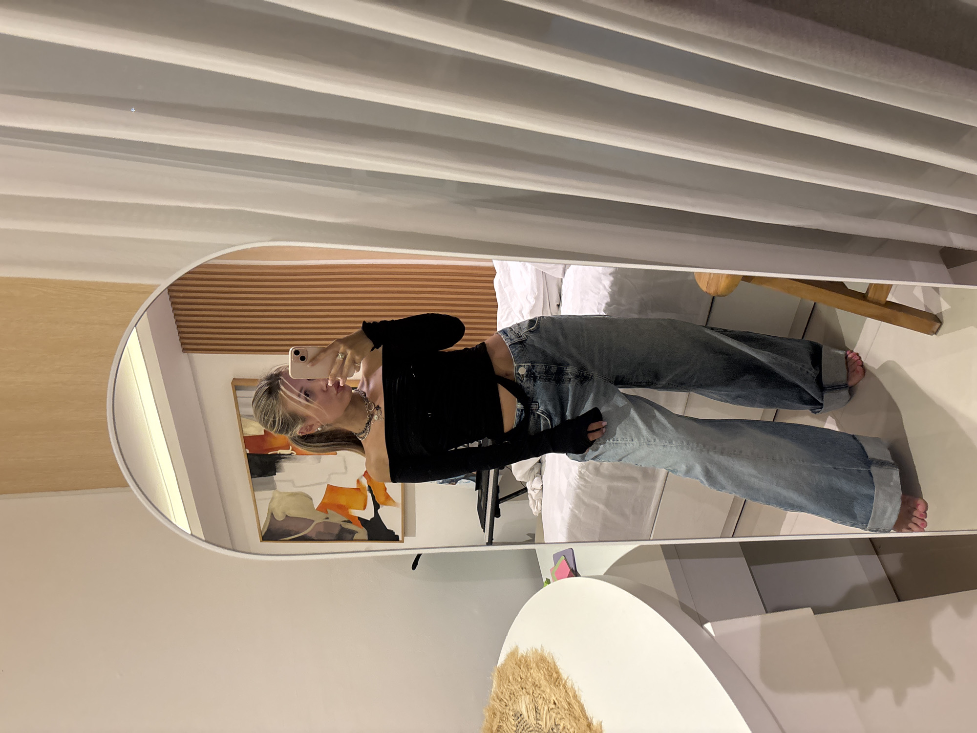 A young woman stands in front of a mirror, taking a selfie. She has blonde hair pulled back into a ponytail and is wearing a black off-the-shoulder long-sleeved top with a criss-cross detail at the midriff and light-wash wide-leg jeans that are cuffed at the bottom. She is accessorized with a chain necklace and several rings. The background shows a bedroom with a bed, a painting on the wall, and sheer white curtains. The lighting is dim, and the overall atmosphere appears to be casual and personal.
