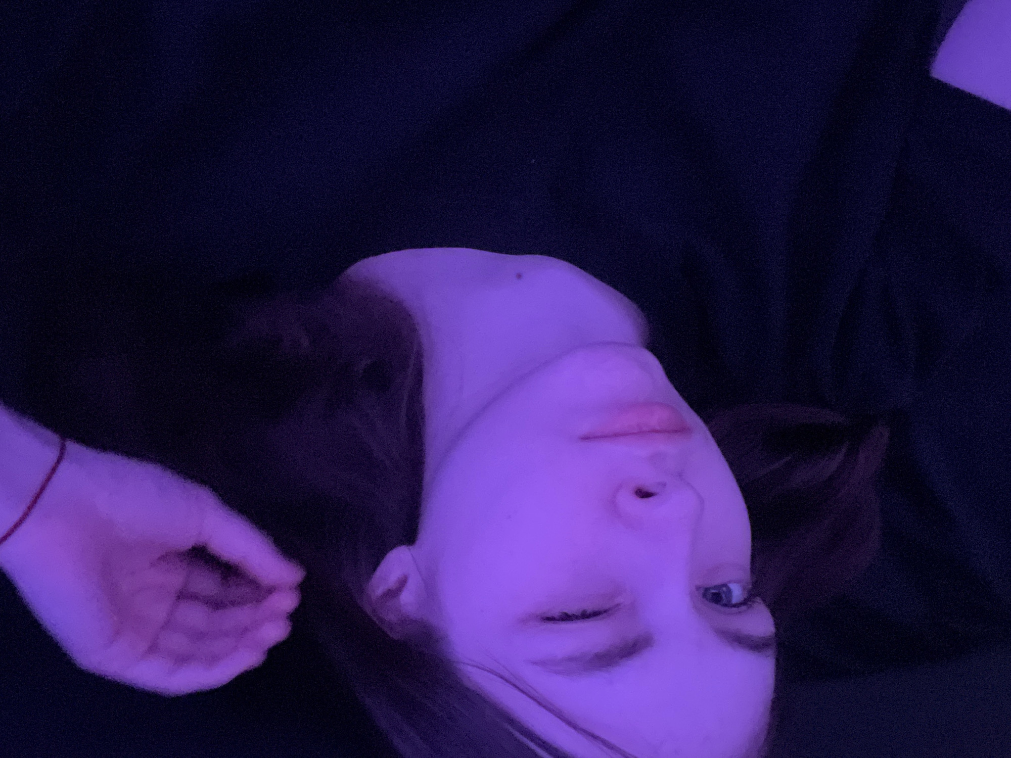 A portrait-oriented, low-angle photograph shows a person lying down, illuminated by a purplish-blue light source. The person's face is tilted to the right. The right eye is closed in a wink, while the left eye is open and appears light blue. The nose is centered in the frame, and the lips are closed. A mole is visible on the neck. The person's left hand is partially visible at the bottom left of the frame, with a red string bracelet around the wrist. The hair is brown. The background is dark and indistinct. The scene does not appear to be in the city of Saint Petersburg, Russia.
