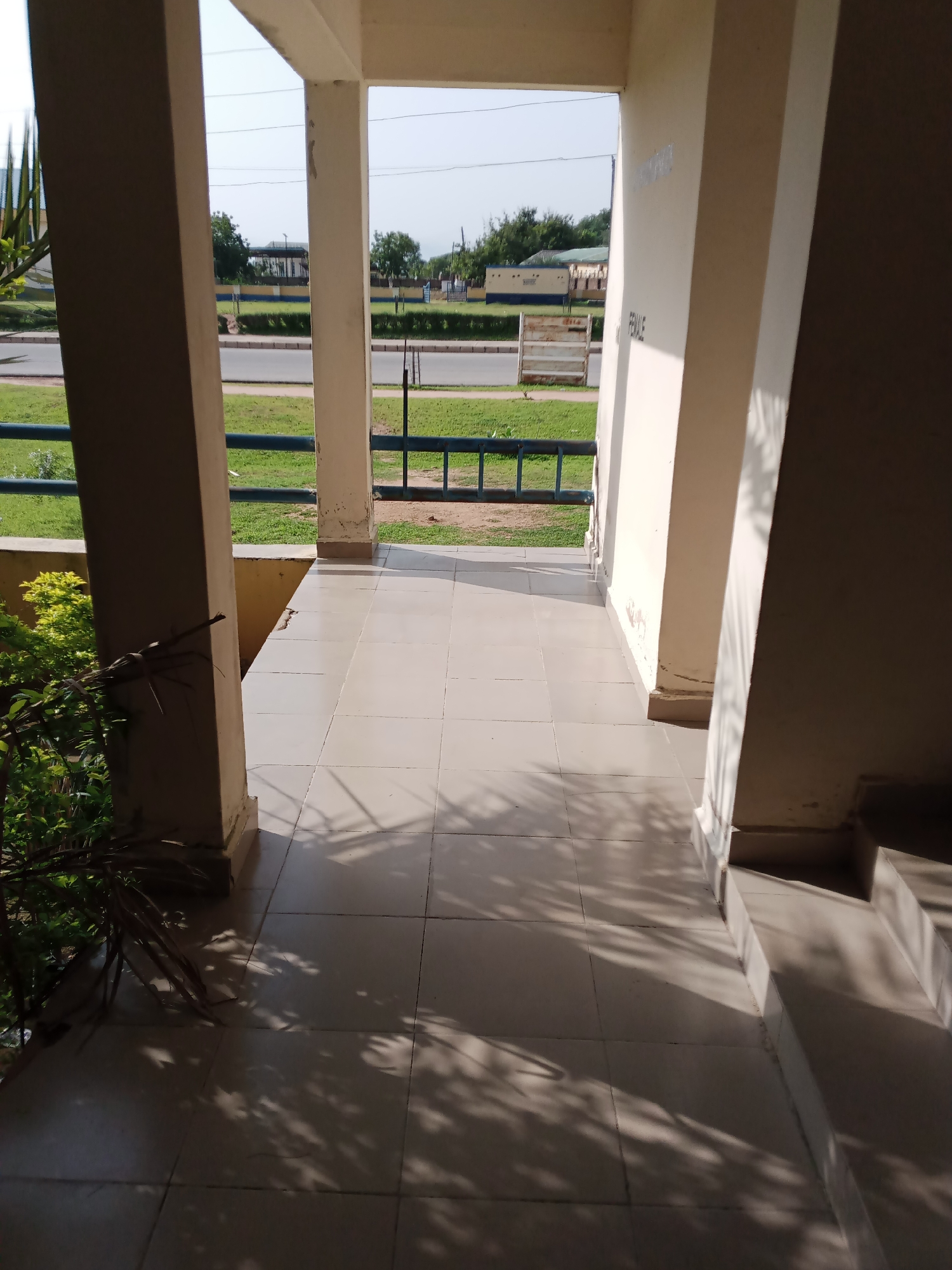 The image depicts an outdoor walkway or veranda in Jalingo, Nigeria, with a tiled floor and overhead cover supported by columns. The walkway leads to an opening revealing a street, green grass, and some buildings in the distance. Shadows of tree leaves are cast on the floor, indicating a sunny day. On the right side of the walkway, there is a wall with some fading text that appears to say "FEMALE". There are also steps visible on the far right, leading downwards. To the left, lush green foliage is present. The overall scene is one of an outdoor public space, likely part of a school or institutional building, given the "FEMALE" signage.