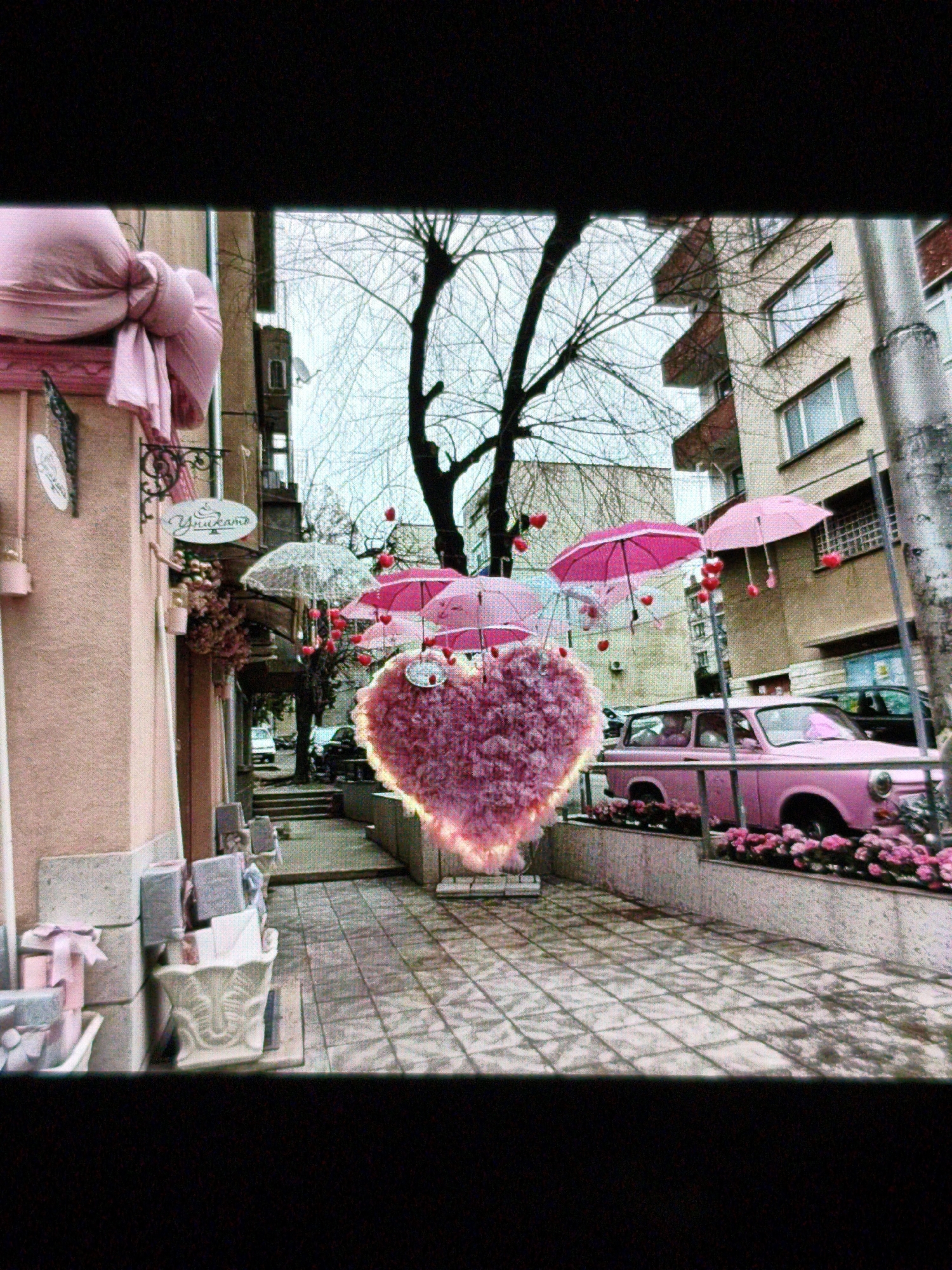The image captures a street scene in Devin, Bulgaria, decorated for Valentine's Day. A large, pink heart-shaped decoration with a glowing aura hangs suspended in the air. Pink umbrellas with dangling red hearts are also suspended. A pink Trabant car sits behind a concrete planter box filled with pink flowers. Buildings line the street, and a tree with bare branches is visible between the decorations. Paved tiles cover the street. A shop or cafe on the left has pink decorations and a sign in Cyrillic. There are no people clearly visible in the image.