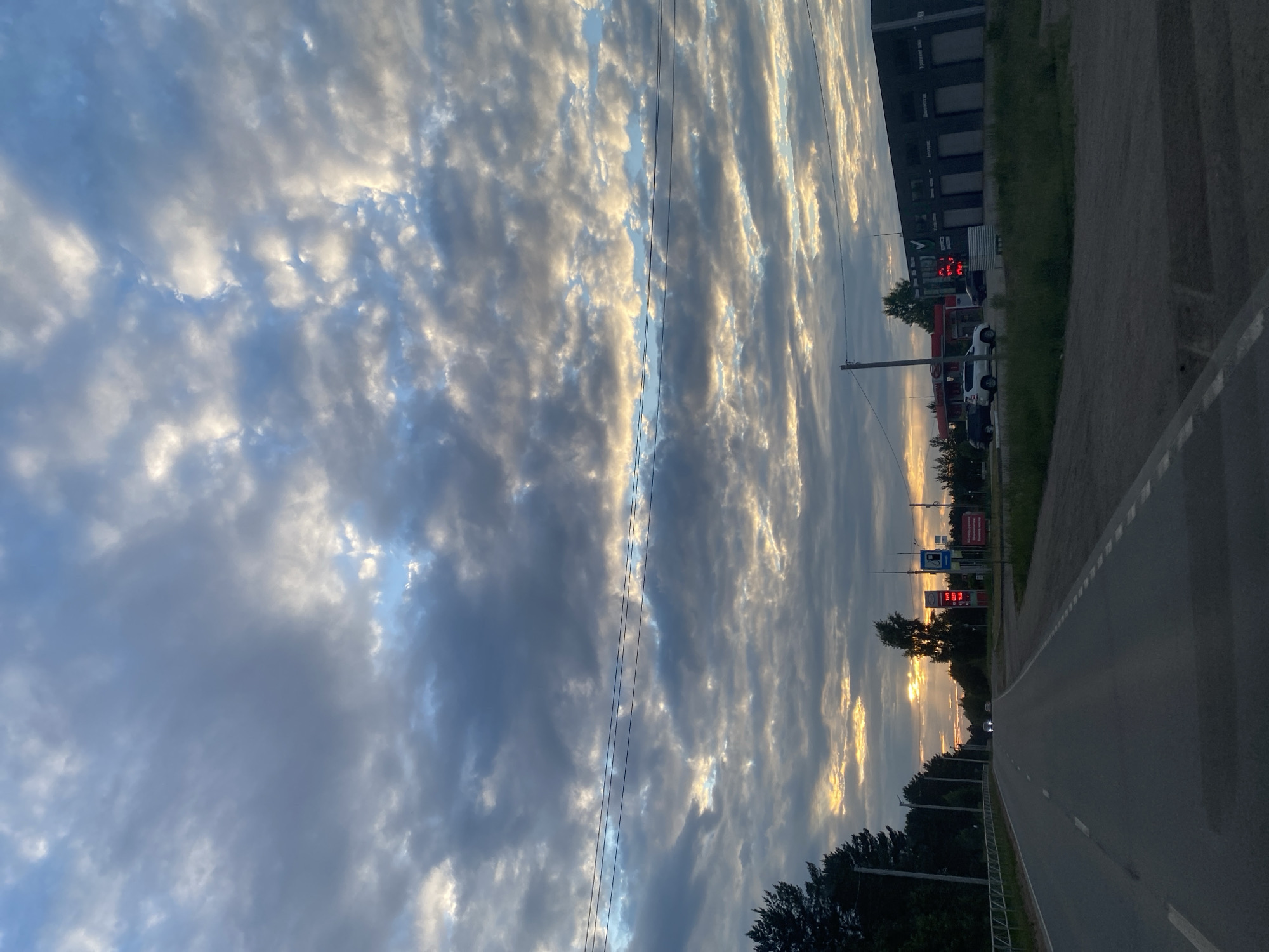 The image captures a scene on a road in Chaikovsky, Russia, during what appears to be sunset or sunrise due to the dramatic lighting of the clouds. The sky is filled with a dramatic display of clouds, with streaks of golden light breaking through the darker, grey clouds, creating a striking contrast. This suggests a dynamic weather pattern, possibly post-storm or approaching weather.
In the mid-ground, there is a gas station with pumps and signage. Some of the signs appear to be displaying prices, suggesting a commercial area. A few vehicles are visible, including a white SUV at the gas station. To the left, a long, dark building with multiple garage doors is visible, possibly a commercial or industrial structure. A power line runs diagonally across the frame, a common sight in such landscapes.
The road itself is paved, with white dashed lines marking the lanes, indicating it is a public road. The road curves slightly to the right as it recedes into the distance. On the right side of the road, a grassy verge and some trees are visible. The overall atmosphere is one of a transitional time of day, with the fading or rising sun casting long shadows and illuminating the sky in a breathtaking manner. The scene evokes a sense of calm, tinged with the dramatic beauty of nature. The time of day is clearly indicated by the low angle of the sun and the colors in the sky, likely evening.