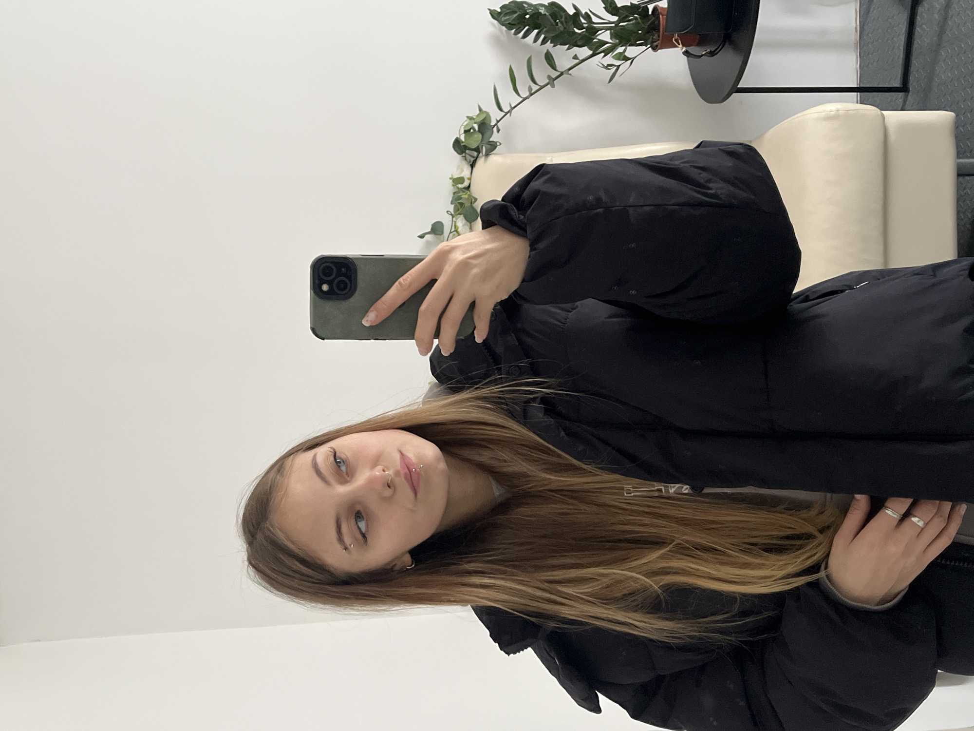 A young woman is taking a selfie indoors. She has long, light brown hair and is wearing a black puffer jacket. She has multiple piercings on her face. She is holding a smartphone with a green case. Behind her is a white wall, a light-colored chair, and a plant on a small table. The location cannot be verified as being in Фоки, Russia.