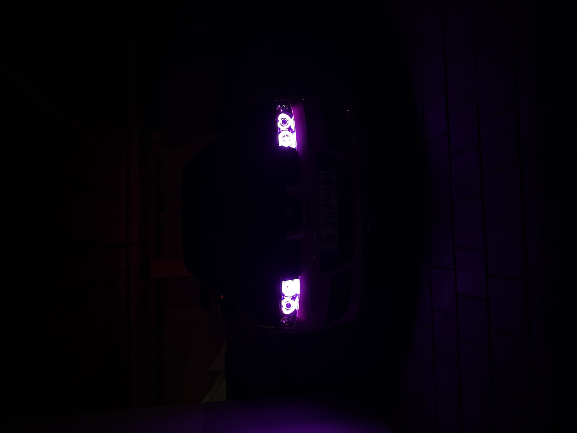 An overhead shot of a car parked in a dimly lit indoor space, possibly a garage. The car's headlights are illuminated with a violet hue, and a license plate with the characters "B 2649 HH" is visible. The surrounding environment is mostly dark, with faint glimpses of wall structures on both sides. The specific location in Varna, Bulgaria, cannot be determined from the image alone.