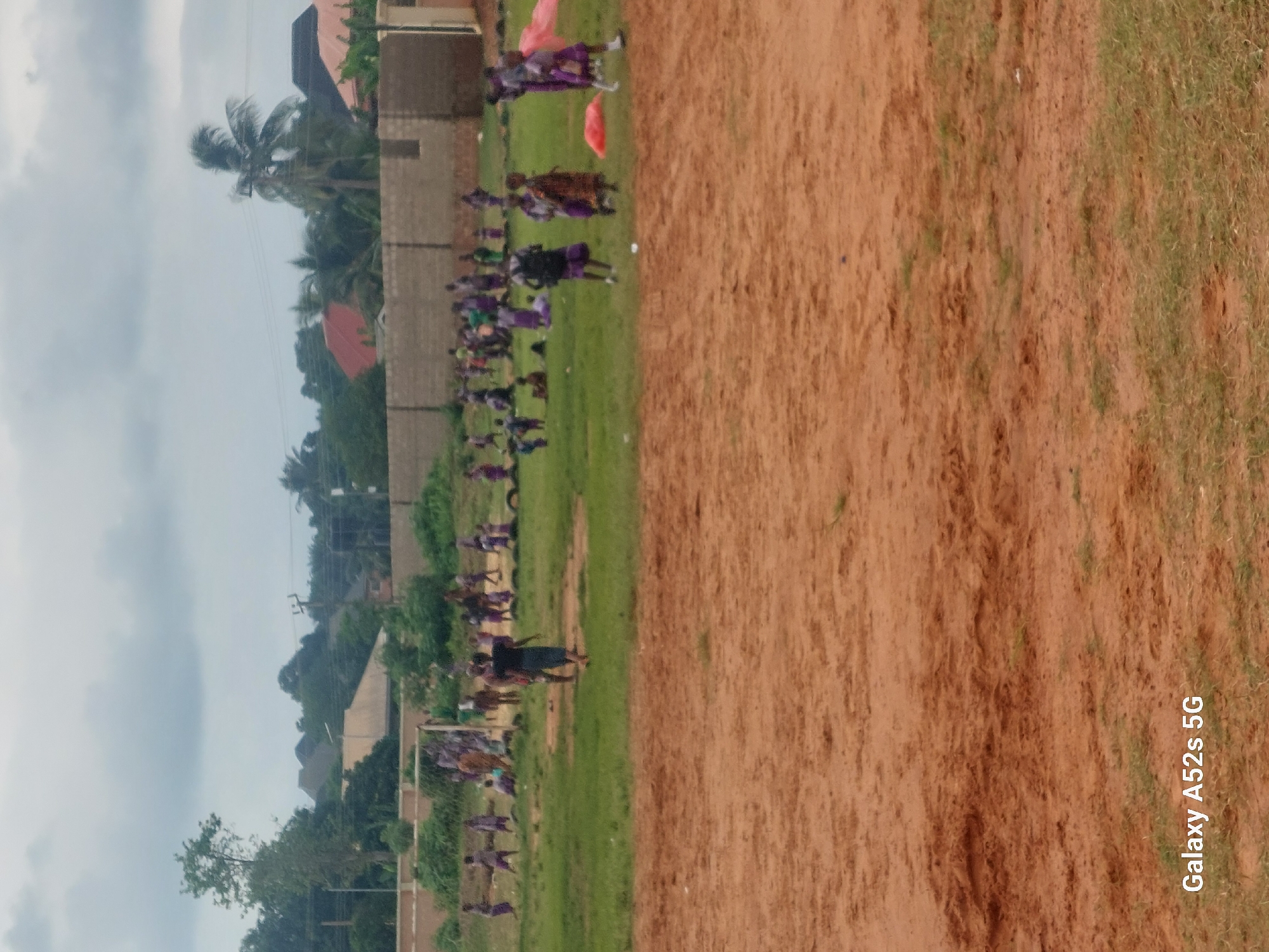 In Umudioka, Nigeria, a group of children are engaged in an outdoor activity on a dirt and grass field. The scene appears to be a school or community gathering, with numerous children scattered across the green grass area. Some children are in motion, while others are gathered in small groups. In the background, a brick wall and fencing delineate the area, with buildings and vegetation visible beyond. The sky is overcast with clouds. The foreground of the image consists of a reddish-brown dirt area.